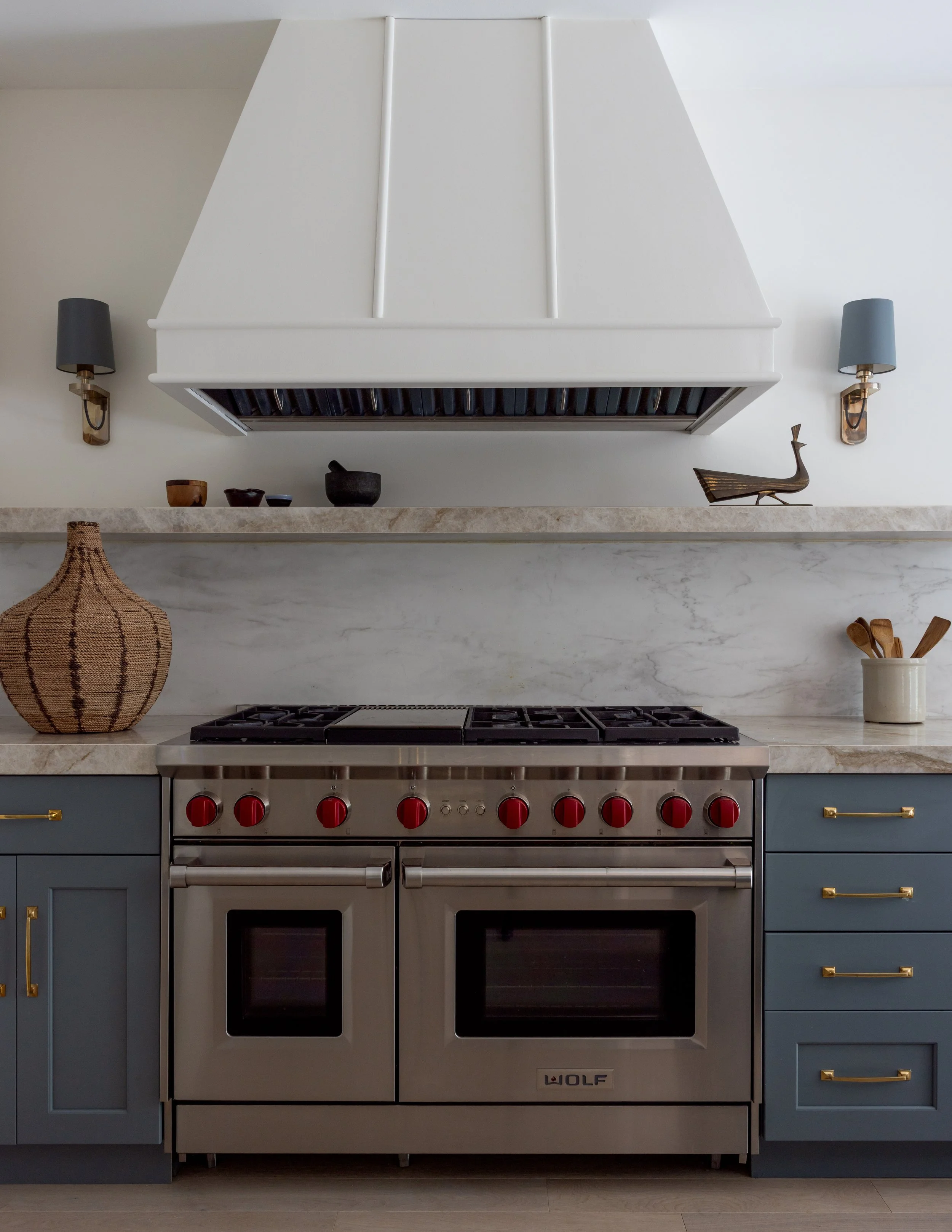 Gorgeous view of the kitchen range with marble shelf, white hood with trim, stainless steel range with red knobs, and blue lower cabinetry with brass pulls inside Imparfait-designed space Crawford