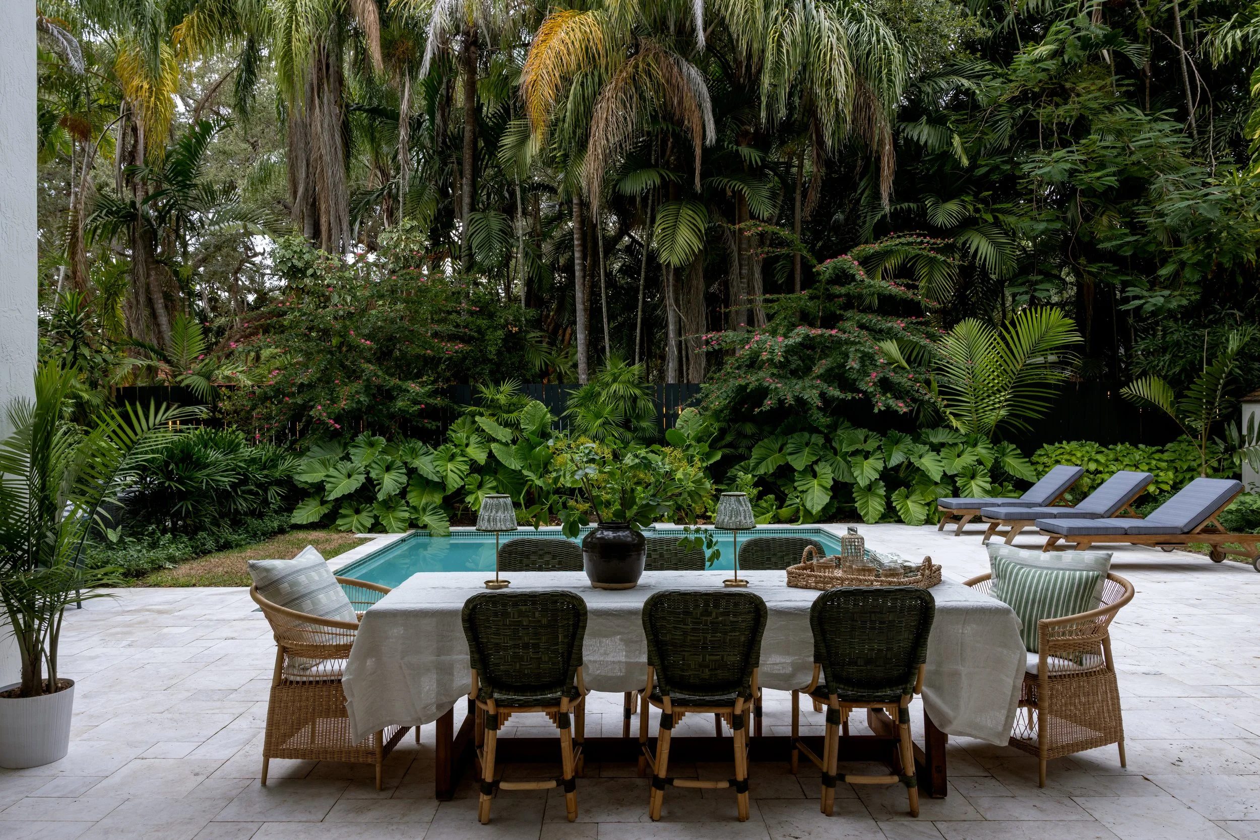 Gorgeous view of the tropical exterior with lush landscaping, a beautiful po a wooden outdoor table with a cream table linen, woven green rattan and bamboo chairs, and curved rattan end chairs with white cushions in Imparfait-designed outdoor space