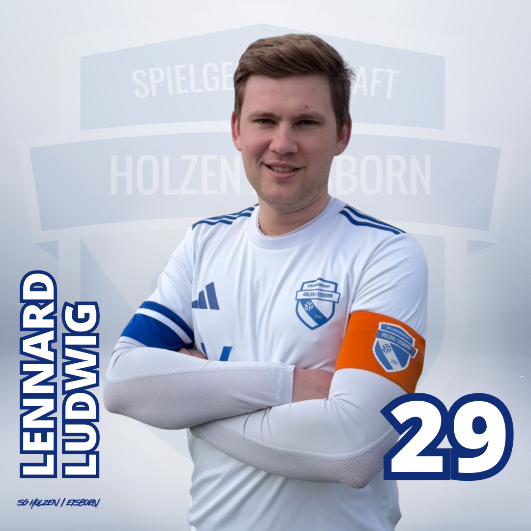 Junger Mann in blauer Fußballtrikot mit Armen vor der Brust, steht vor einem weißen Hintergrund mit einem halbtransparenten Wappen des Holstein Kiel Fußballvereins.