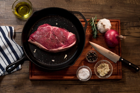 Raw steak in a cast iron skillet on a wooden cutting board surrounded by garlic, red onion, black pepper, salt, and a knife, with olive oil and a striped cloth nearby.