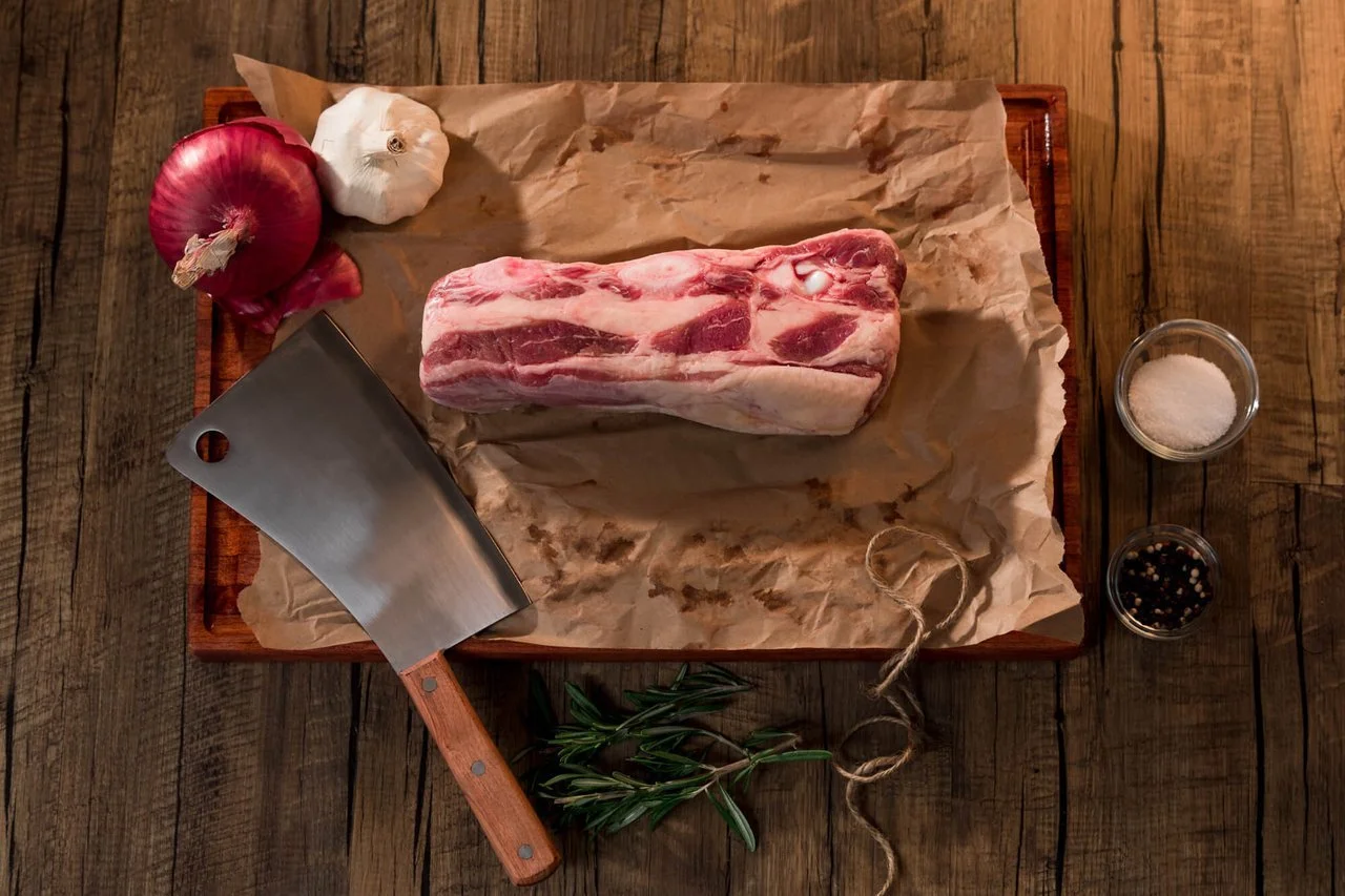 Raw pork shoulder on a wooden cutting board, surrounded by garlic, onion, salt, pepper, and rosemary with a meat cleaver on the side.