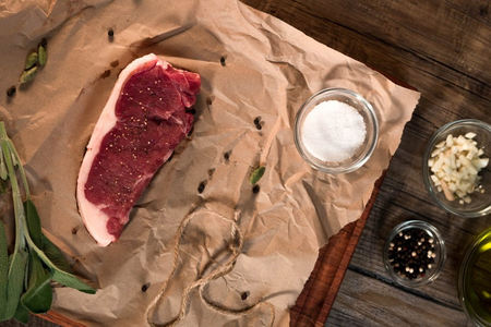 Raw beef steak on brown paper with salt, minced garlic, black pepper, and olive oil nearby.