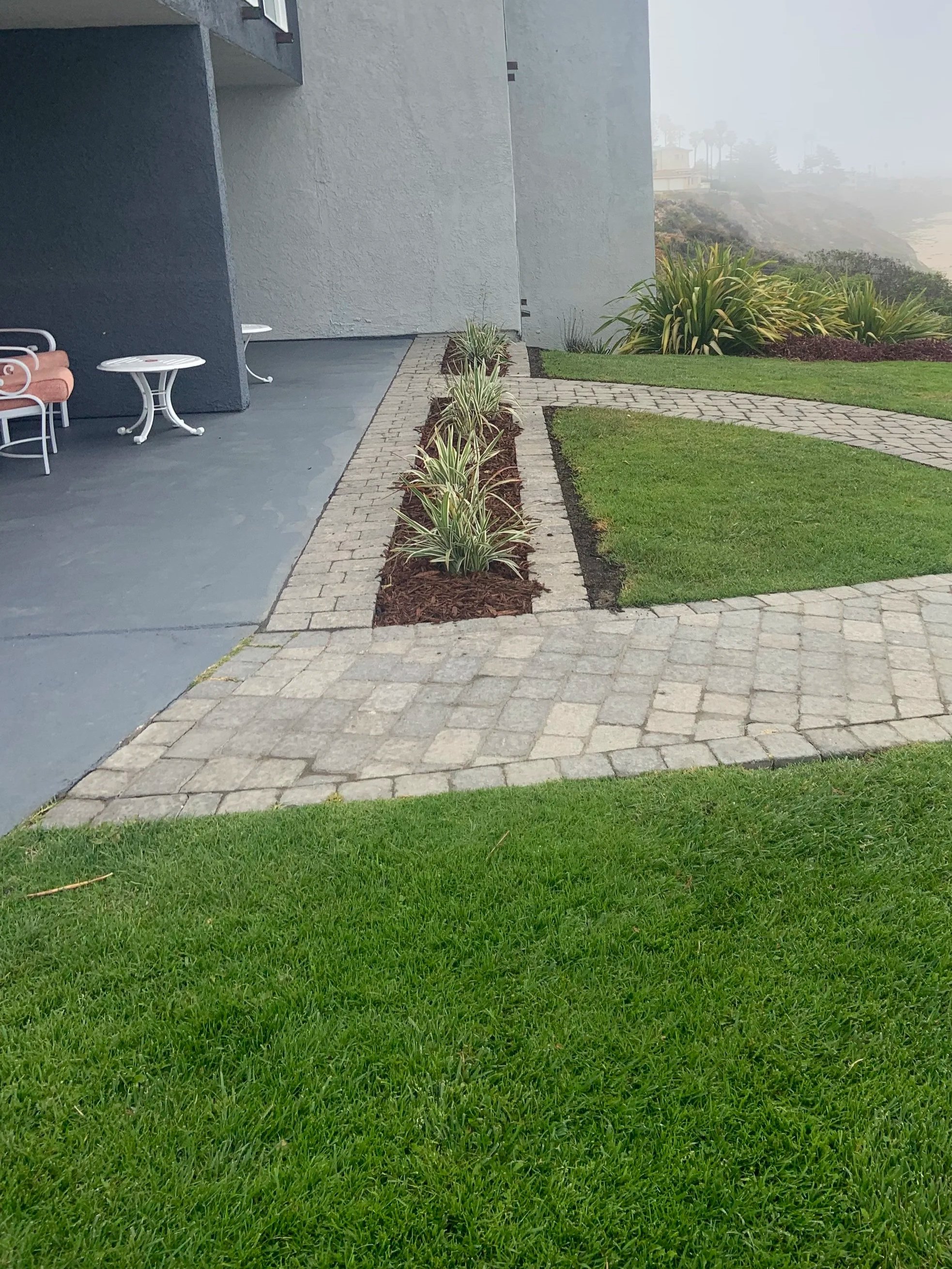 Residential outdoor patio area with small white tables and chairs, a paved walkway, freshly planted flowers, and a grassy lawn near a foggy beach coast.