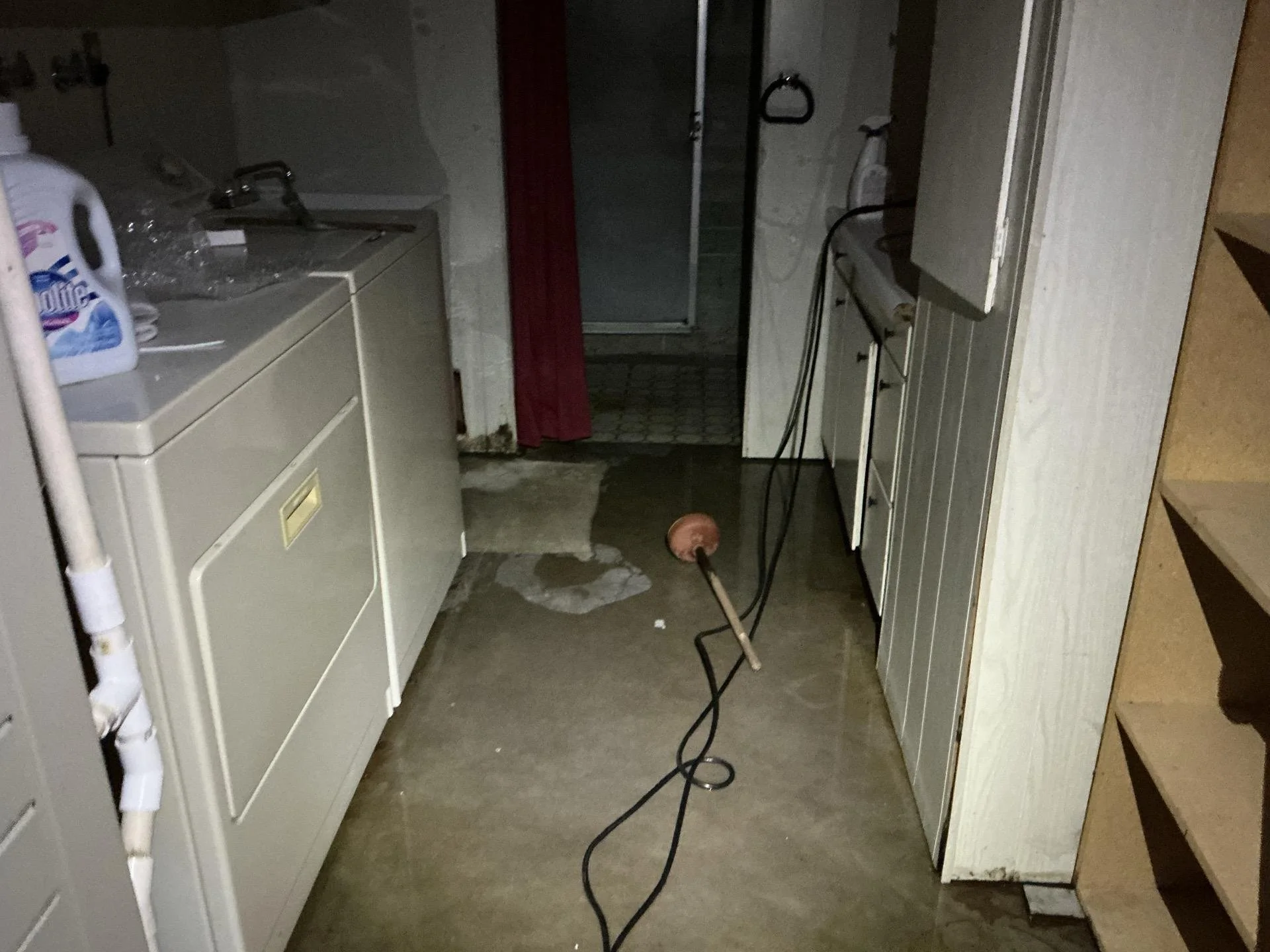 A dimly lit utility or laundry room with a washing machine on the left, a sink, and cabinets on the right. The floor has water damage or a stain, and there is a bent metal pipe or tool on the floor. A red curtain or fixture is visible near a shower or doorway at the back.