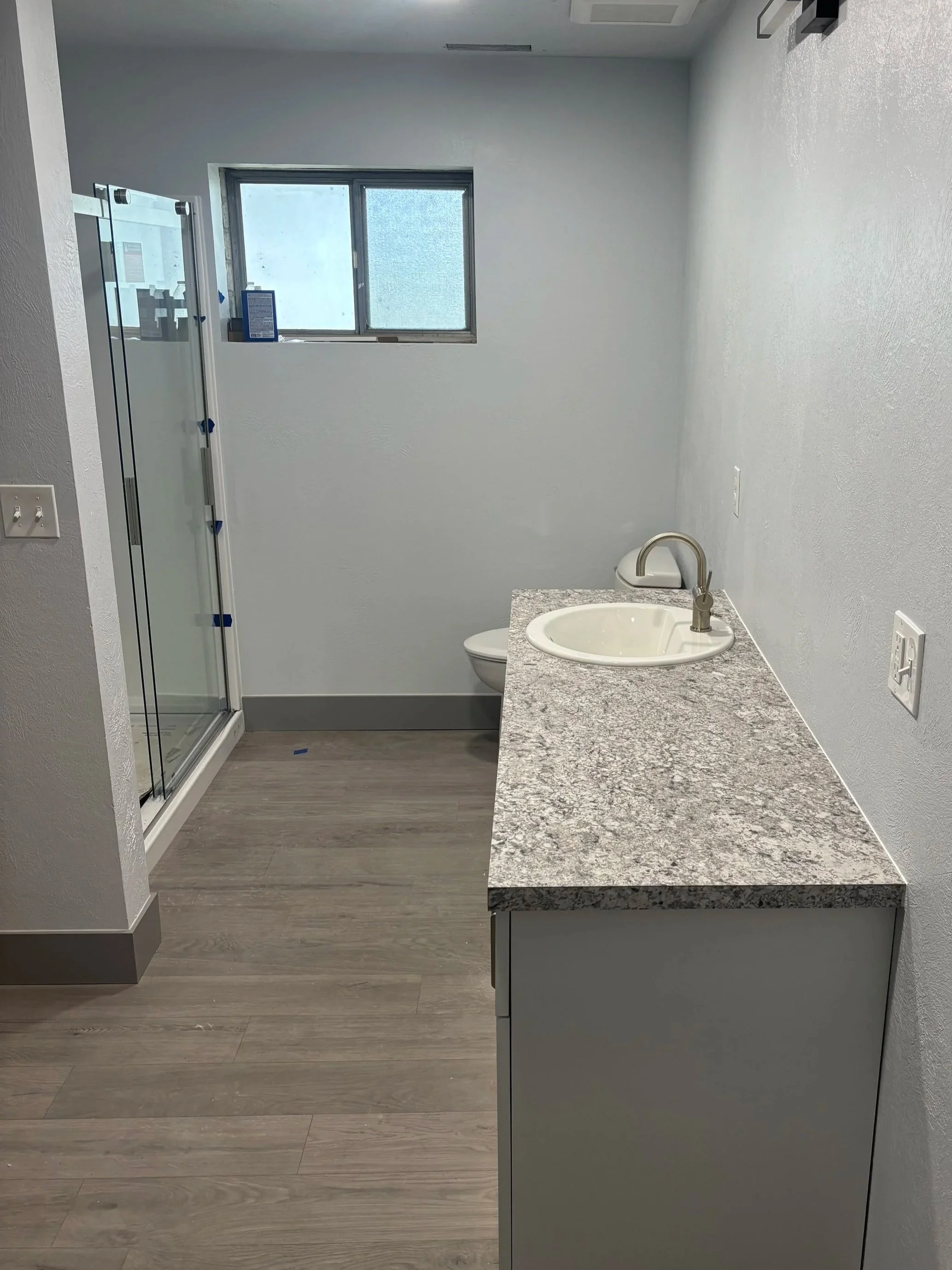A bathroom with a granite countertop, white sink, and silver faucet. To the left, there is a shower with glass doors and blue tape. A small window with frosted glass is above the toilet. The floor has gray wood-like tiles, and the walls are painted white.
