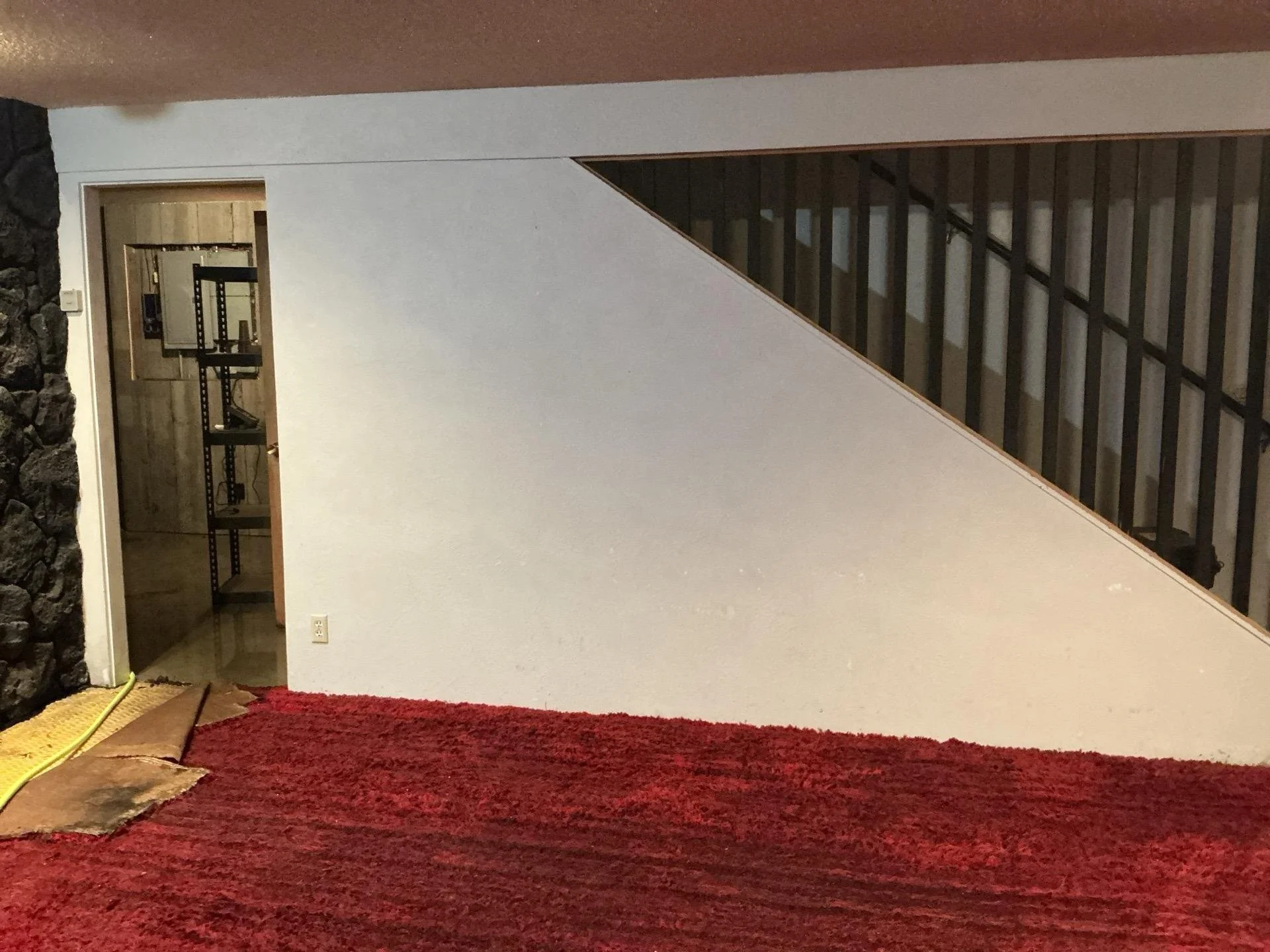 View of a staircase with black railing, a white wall, a doorway revealing a room with shelves, a red carpeted floor, and a stone wall on the left. 