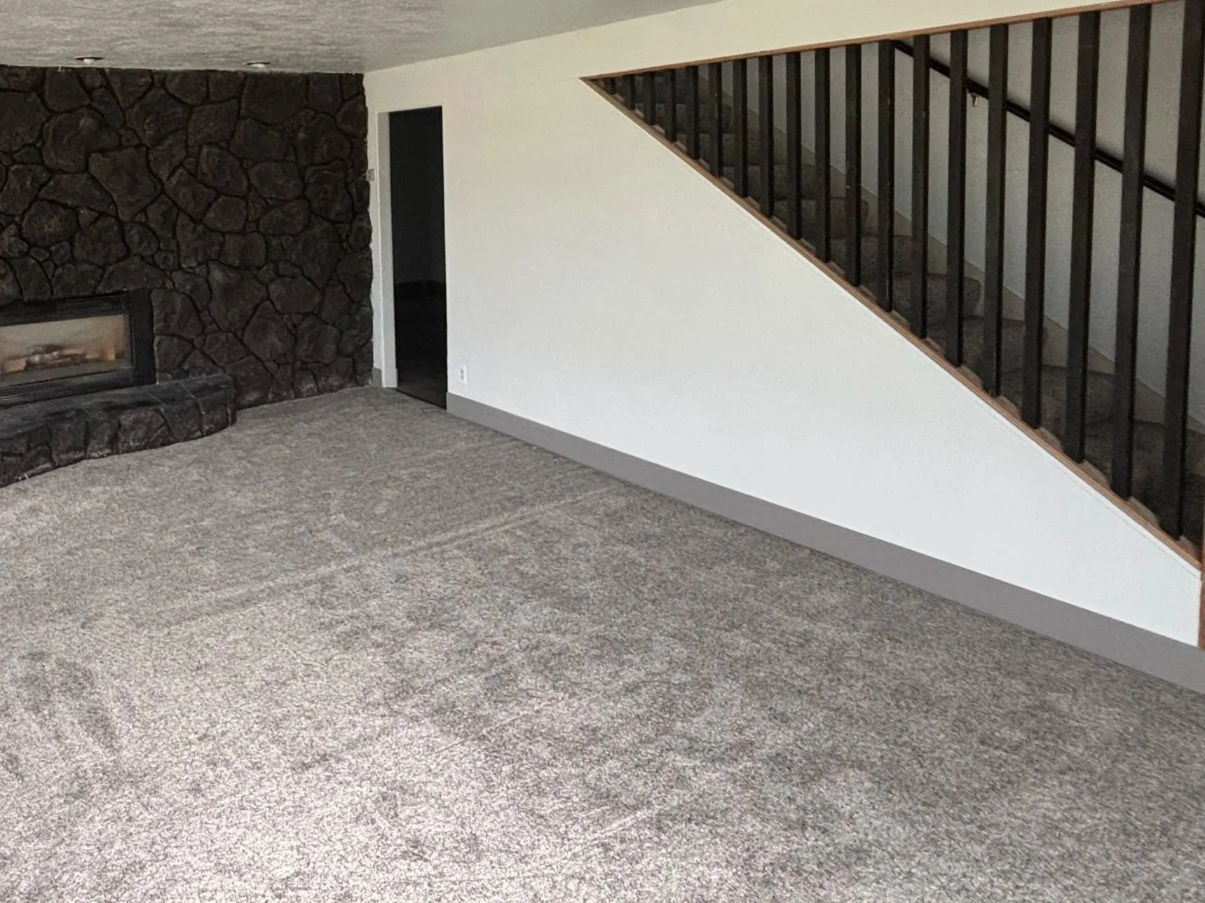 Interior of a living room with a fireplace, beige carpet, and a staircase with black railings against a white wall.