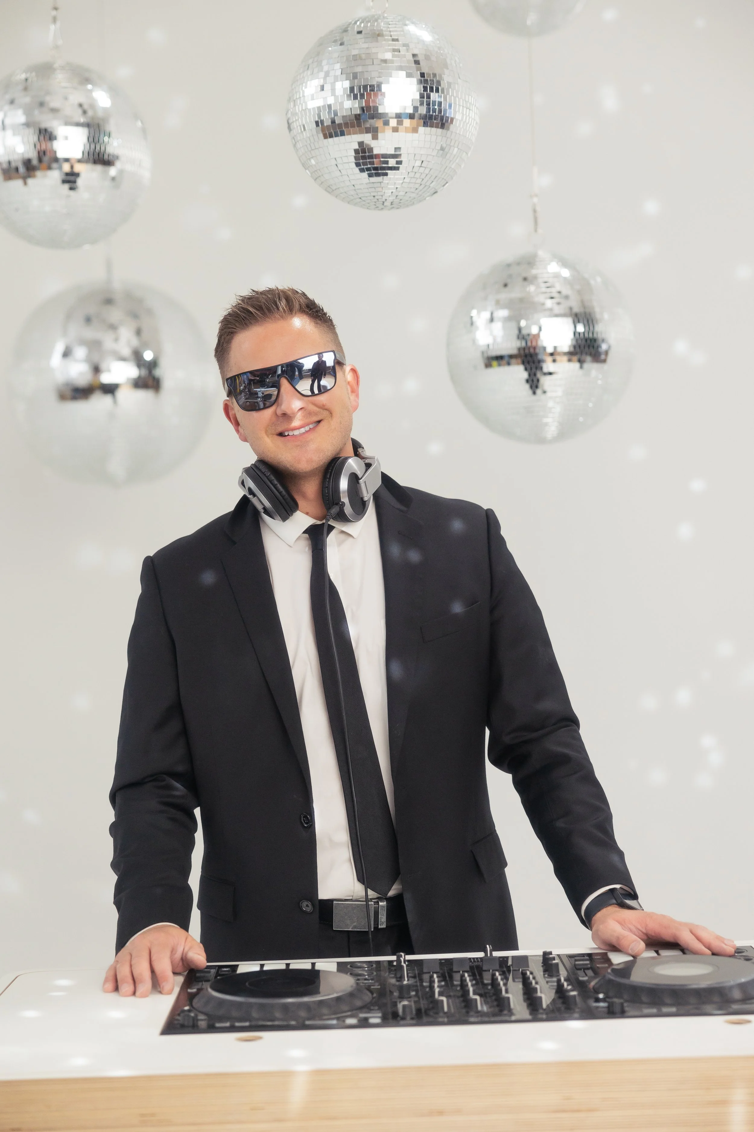 A man in a black suit, white shirt, and black tie, wearing sunglasses and headphones around his neck, standing behind a DJ turntable with disco balls hanging in the background.