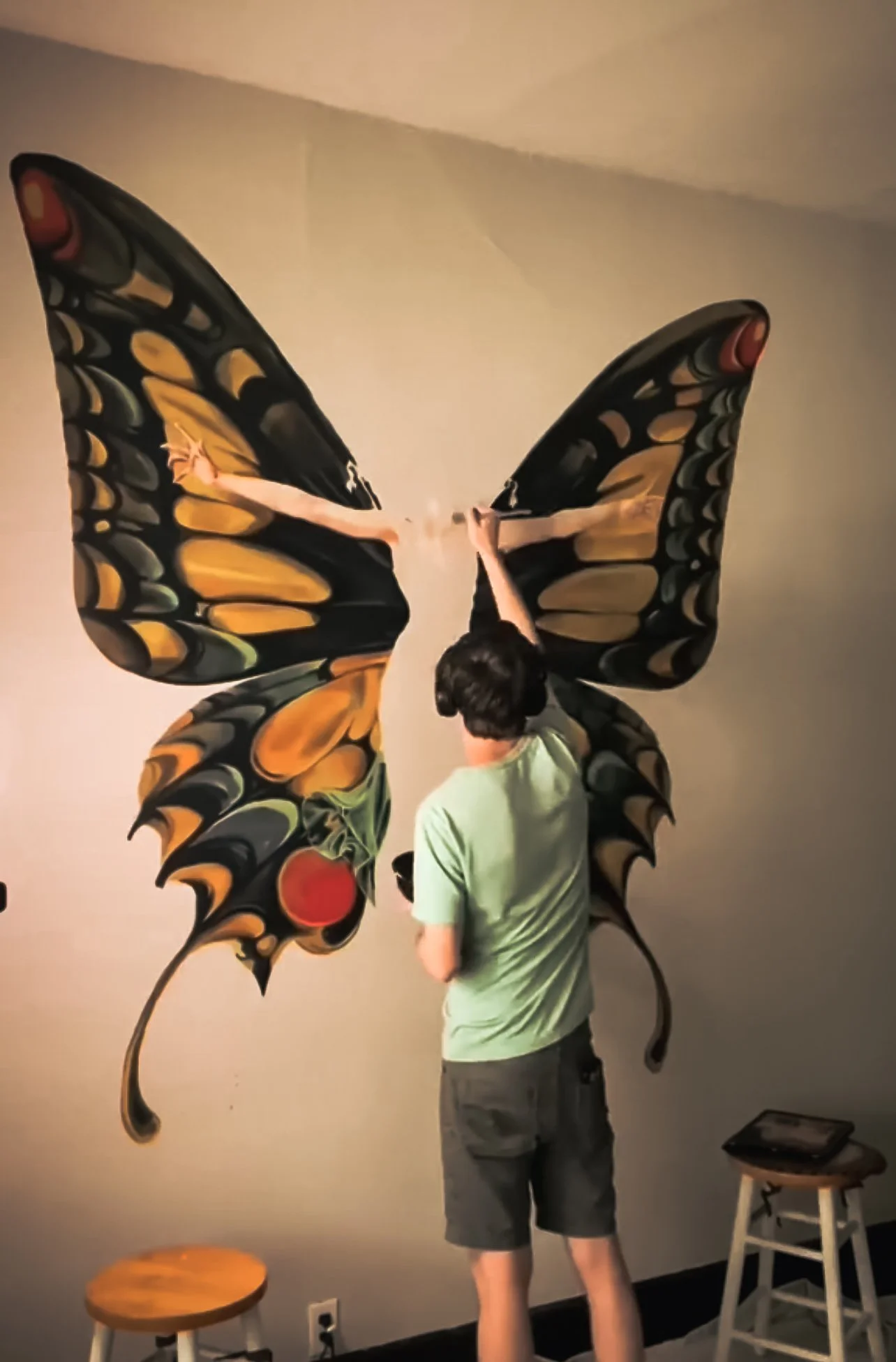 A person stands in front of a large butterfly mural on a wall, painting or decorating its wing with a brush. The butterfly is black with orange and yellow patterns and red spots. There is a small wooden stool and a chair nearby in the room.