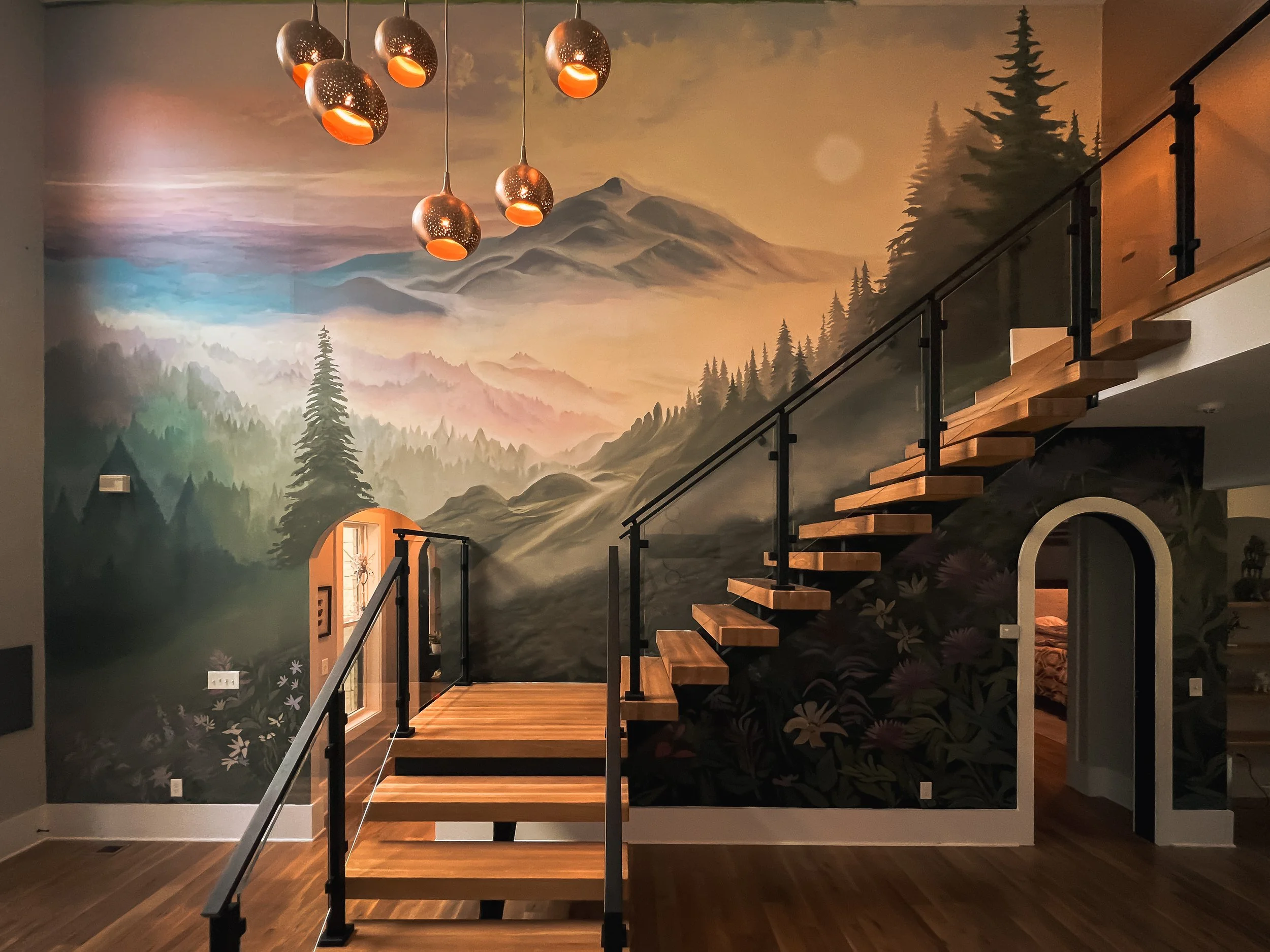 Interior of a house with a staircase and a wall mural of a mountain landscape with trees and mountains, and a painted sky with clouds. Several pendant lights hang from the ceiling.