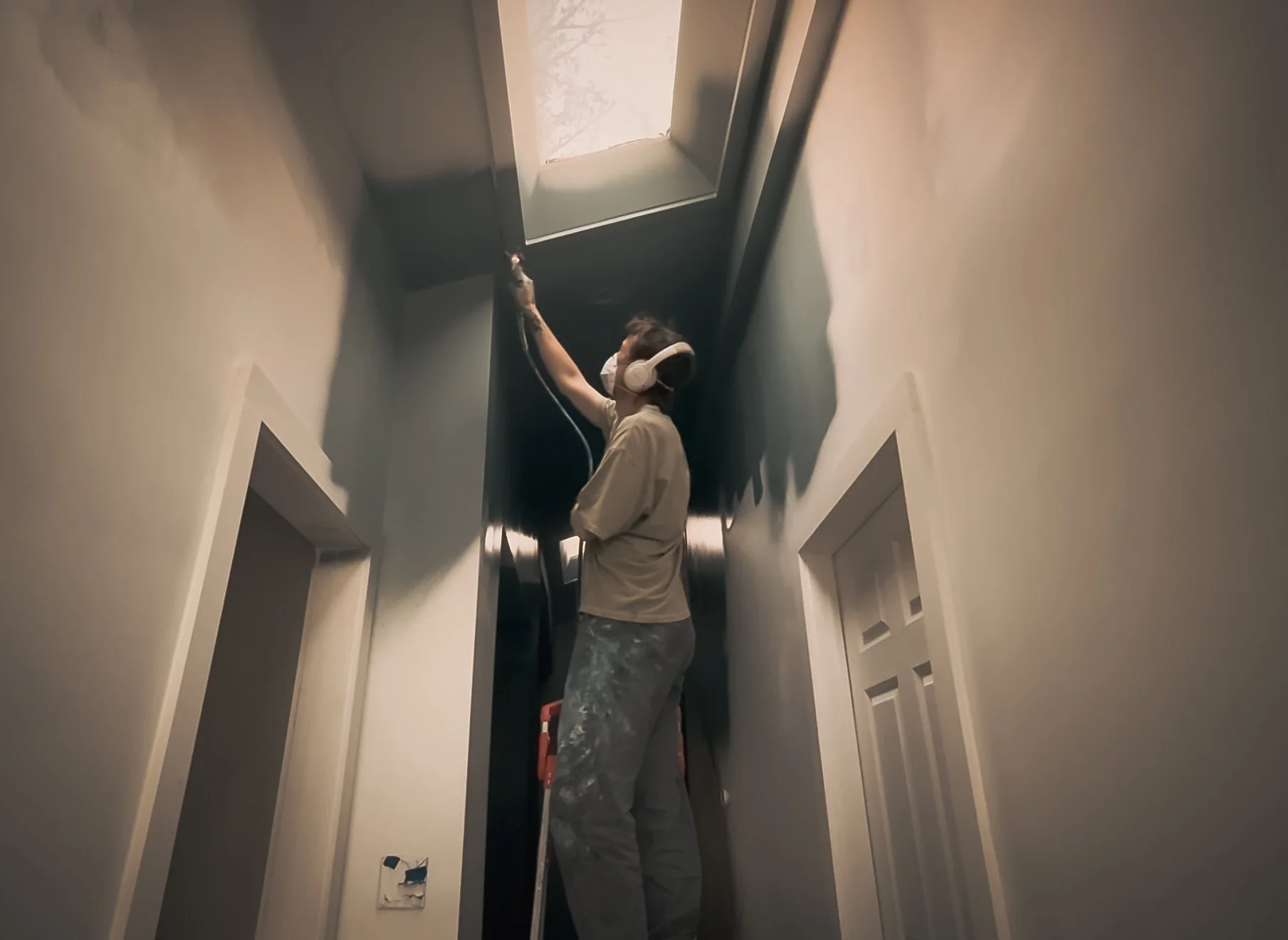 Person wearing a face mask and headphones standing on a ladder, working on ceiling or smoke detector in a narrow hallway.