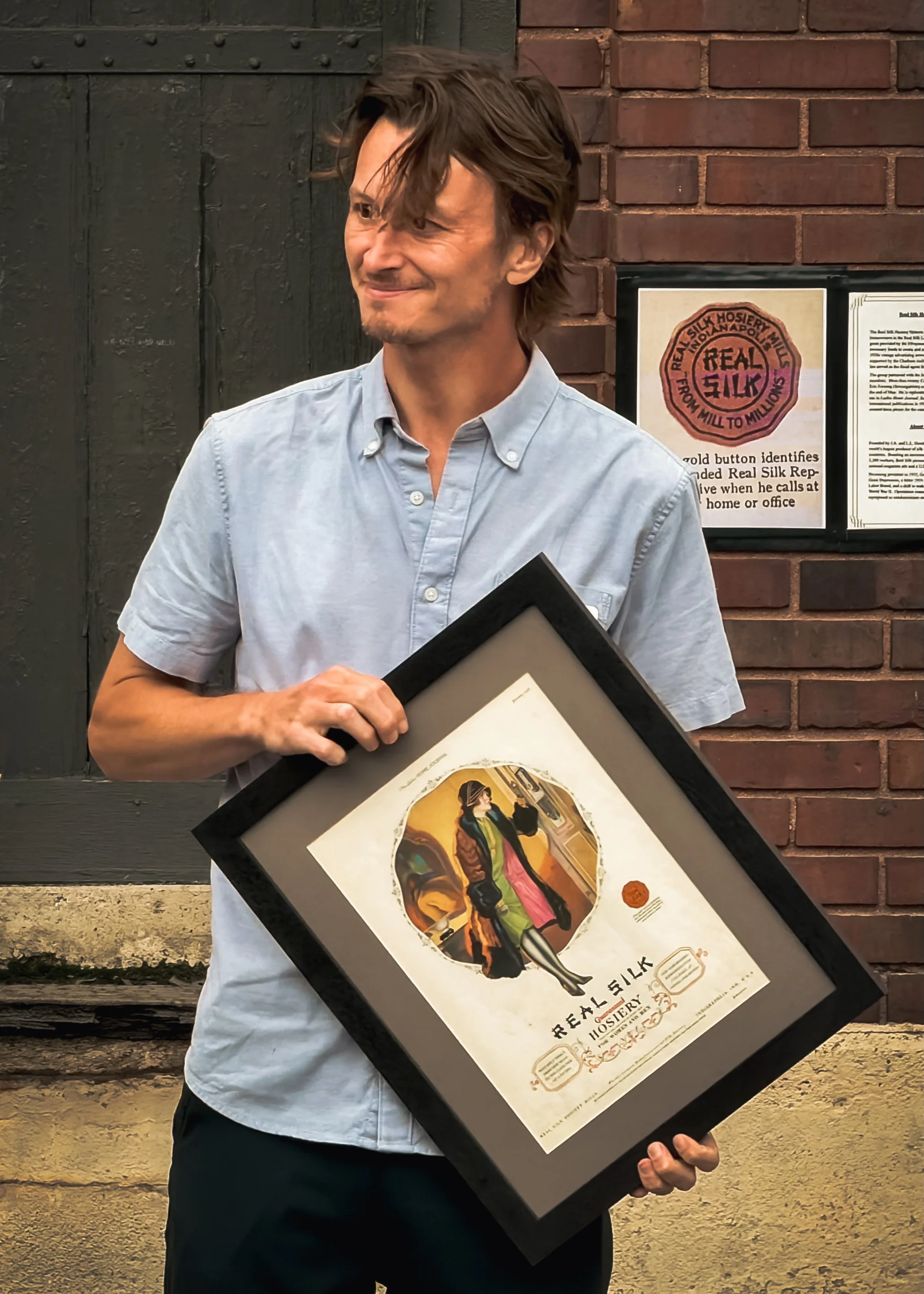 A man in a light blue button-up shirt holding a framed poster in front of a brick wall and black door. The poster features artwork and text related to a whiskey brand, Real Silk Hosiery Mill, with a circular logo and colorful illustration.