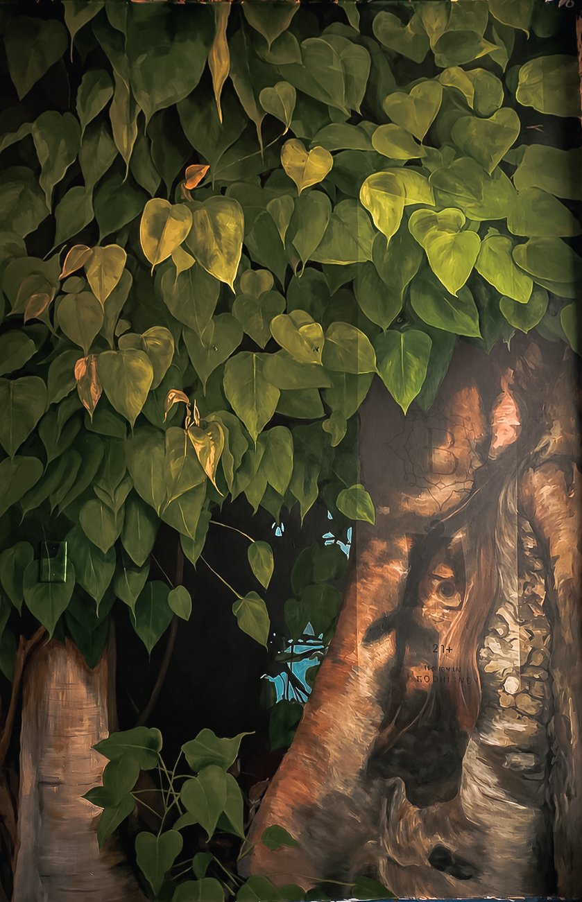 Bodhi Tree & Foliage Interior Mural Detailed interior nature mural featuring dense green foliage, climbing leaves, and a large textured tree trunk painted across the wall.