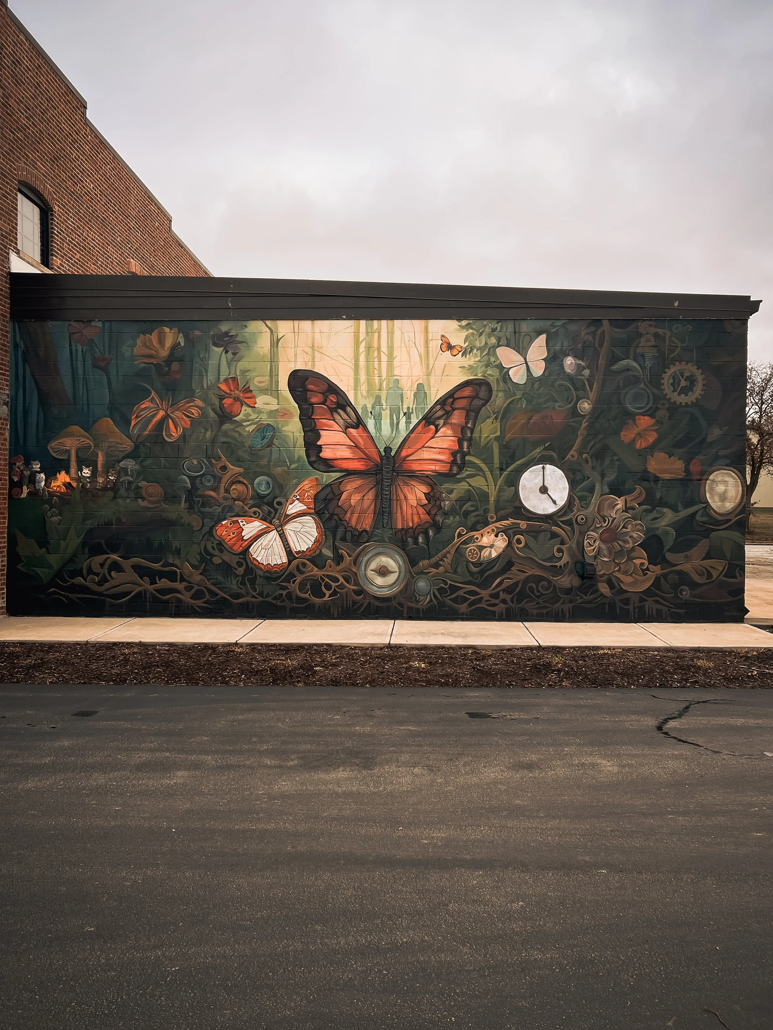 A colorful mural painted on the side of a brick building, featuring large butterflies, gears, and abstract designs. The building has a small window with a curved top, and there is a concrete sidewalk and some freshly landscaped soil in front of it.