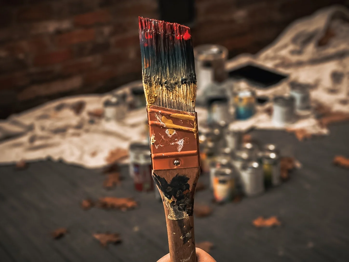 Close-up of a paintbrush held up with a blurry background featuring a table with scattered paint cans and a cloth, and a brick wall behind.