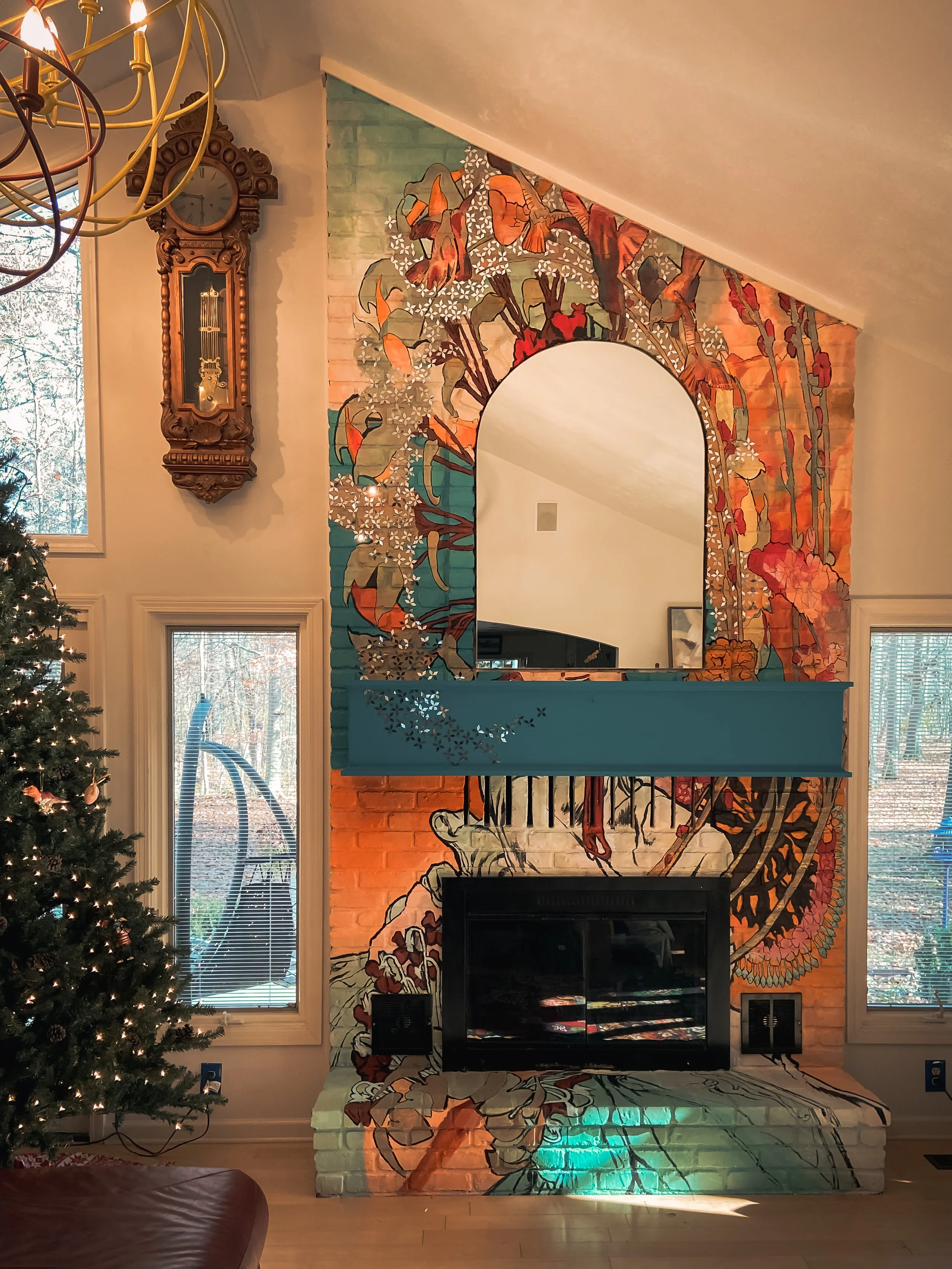 Living room with a Christmas tree on the left, a colorful mural on the wall behind a black fireplace, with a mirror reflecting part of the mural, and two large windows showing trees outside.