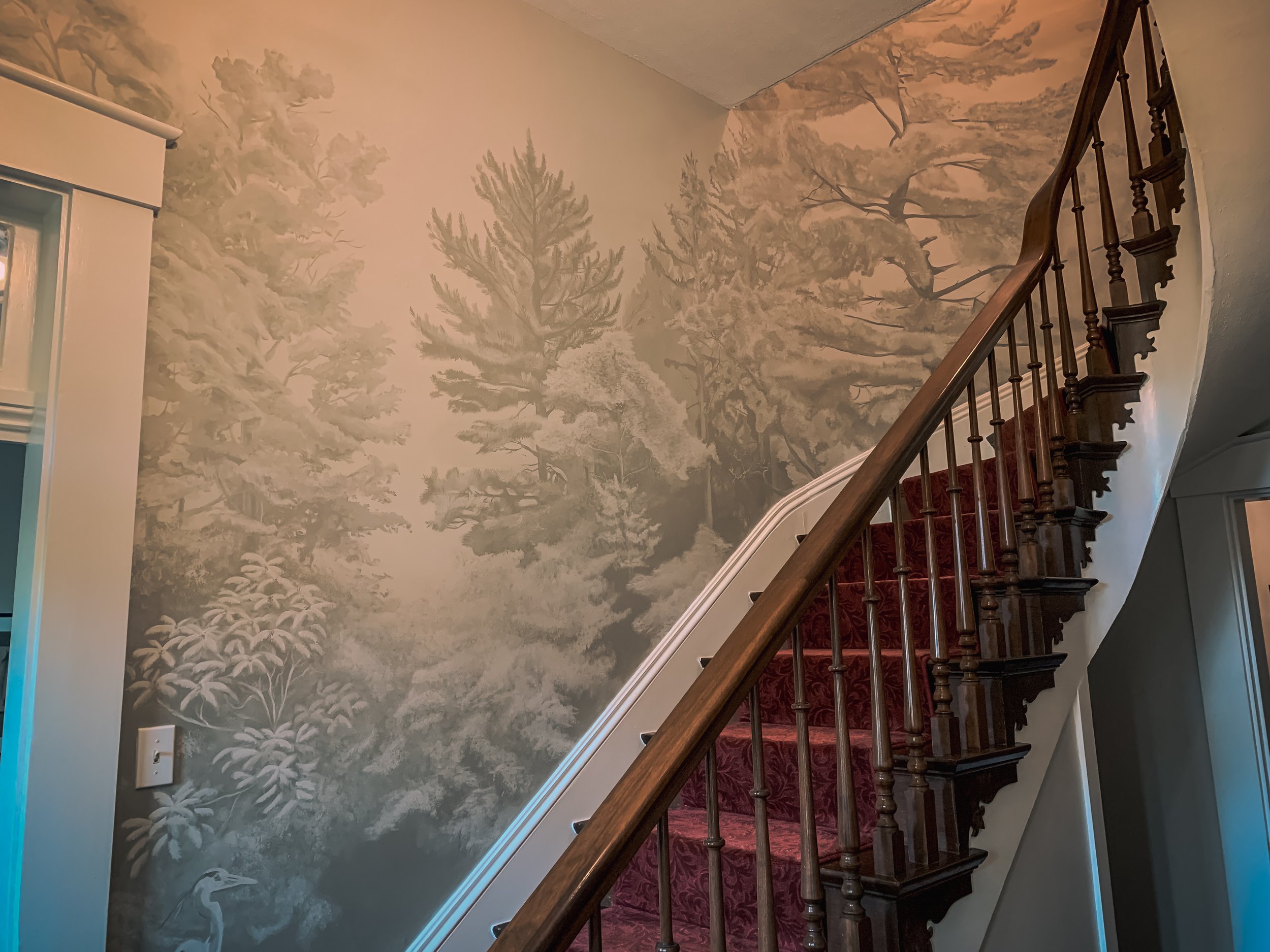 Monochromatic woodland mural integrated around a residential staircase, featuring layered trees, dense foliage, and a heron.