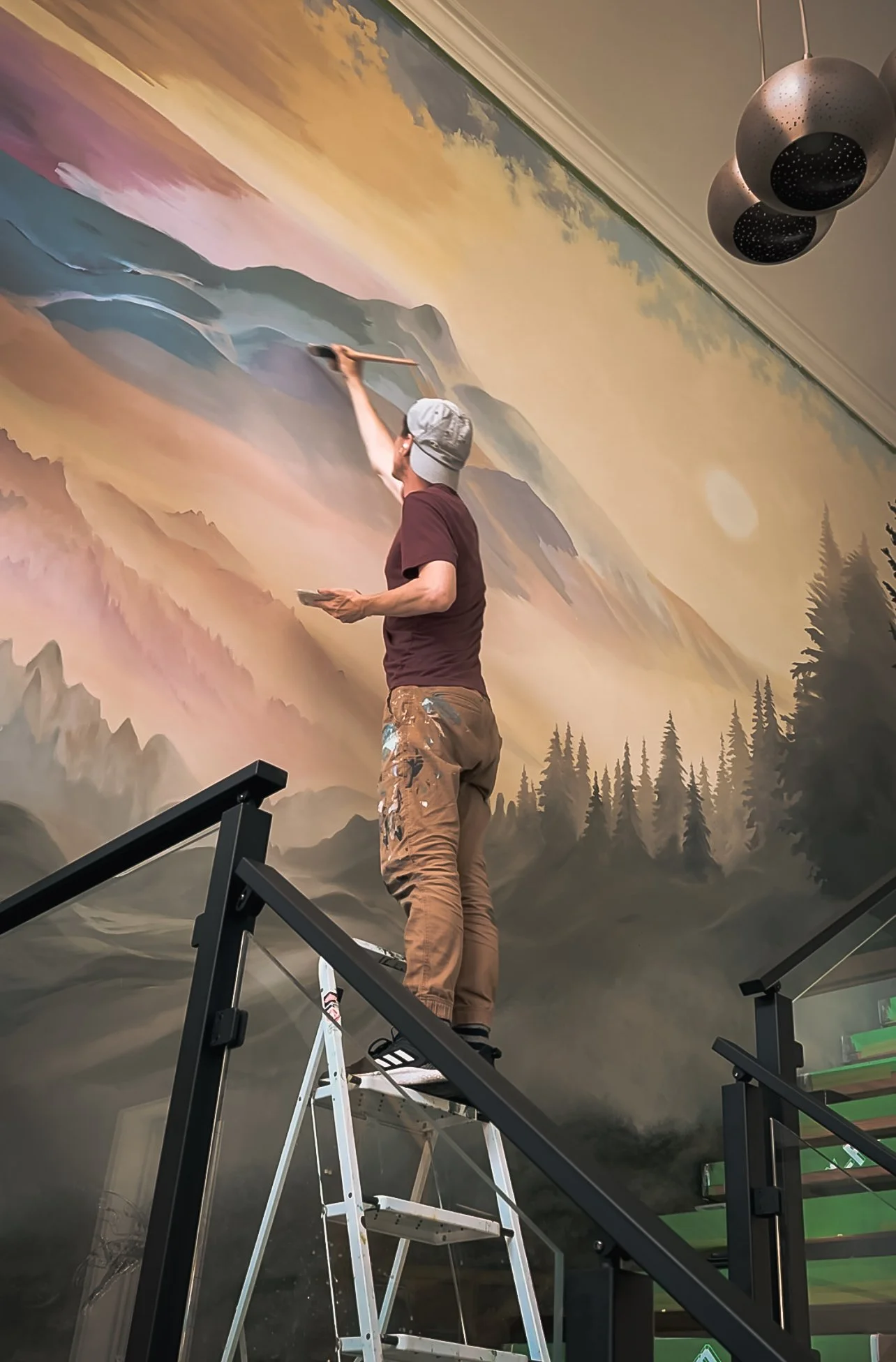 A man painting a detailed landscape mural on a high wall, standing on a ladder. The mural depicts mountains, trees, and a sunset sky.