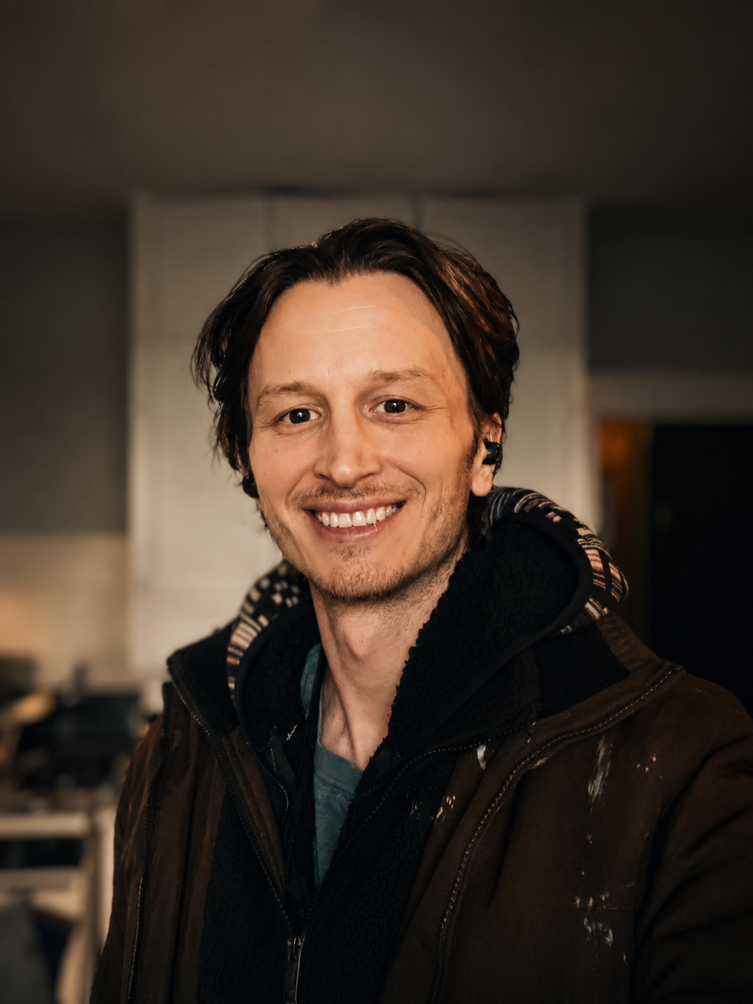 A smiling young man with dark hair, wearing a black hoodie and jacket, standing indoors.