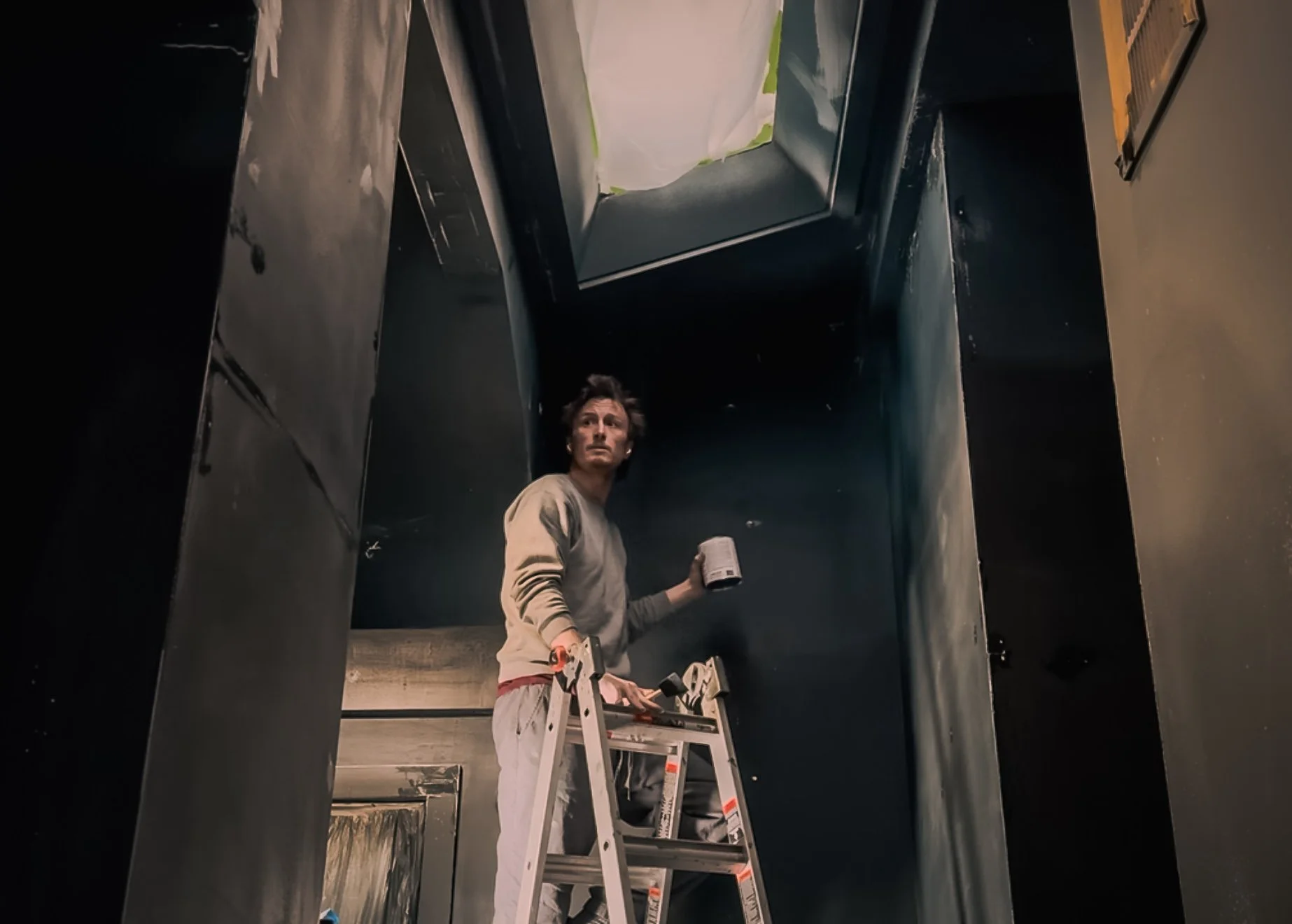 A man on a step ladder working on the ceiling of a dark industrial room, holding a container of paint.