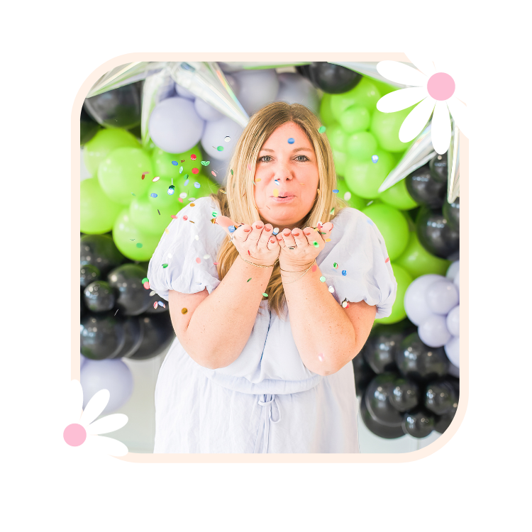 Woman blowing colorful confetti amid green, black, and gray balloons with floral decorations.