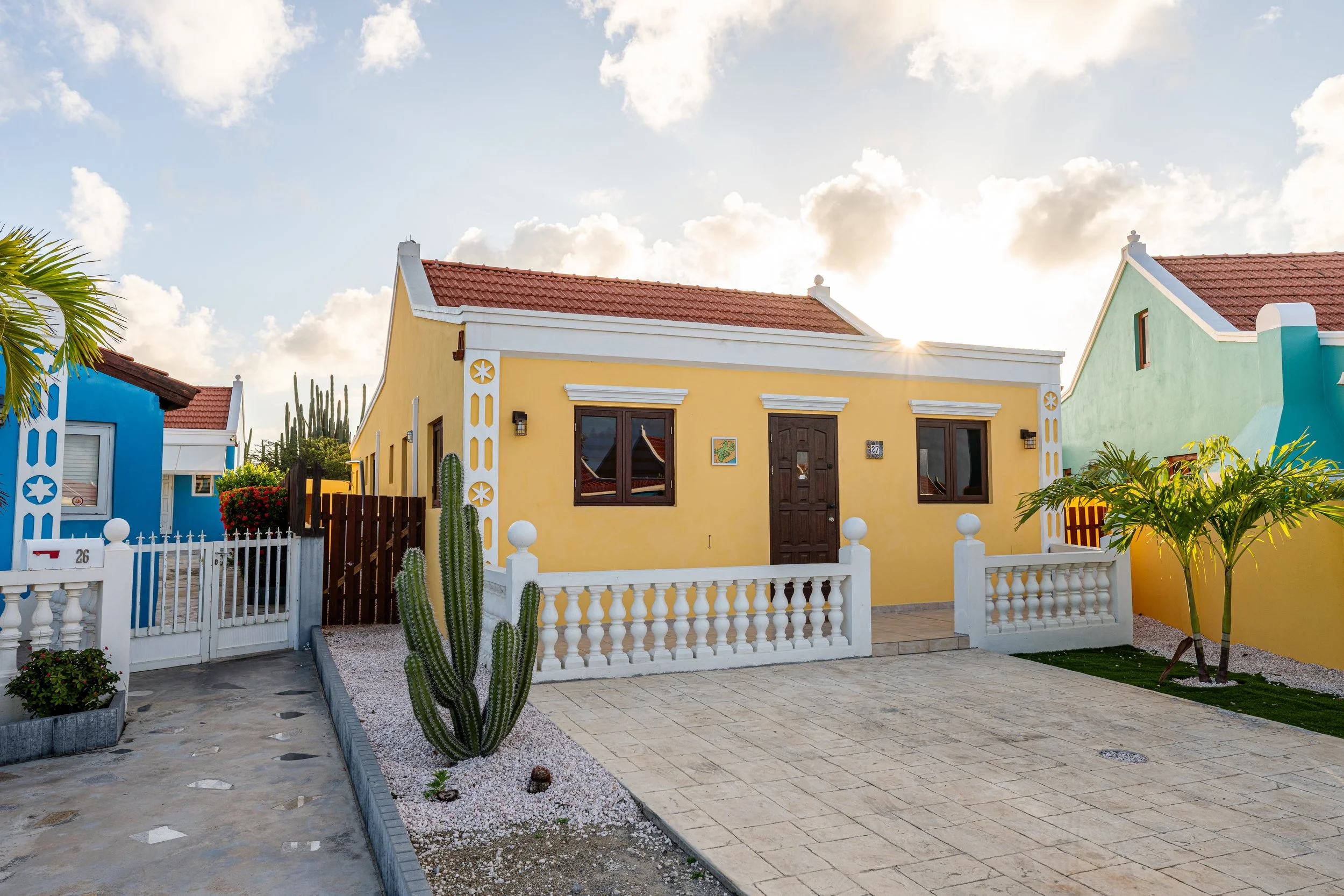 Colorful house with yellow walls, white decorative trim, brown door, and black windows, with desert plants and trees in the front yard under a partly cloudy sky.