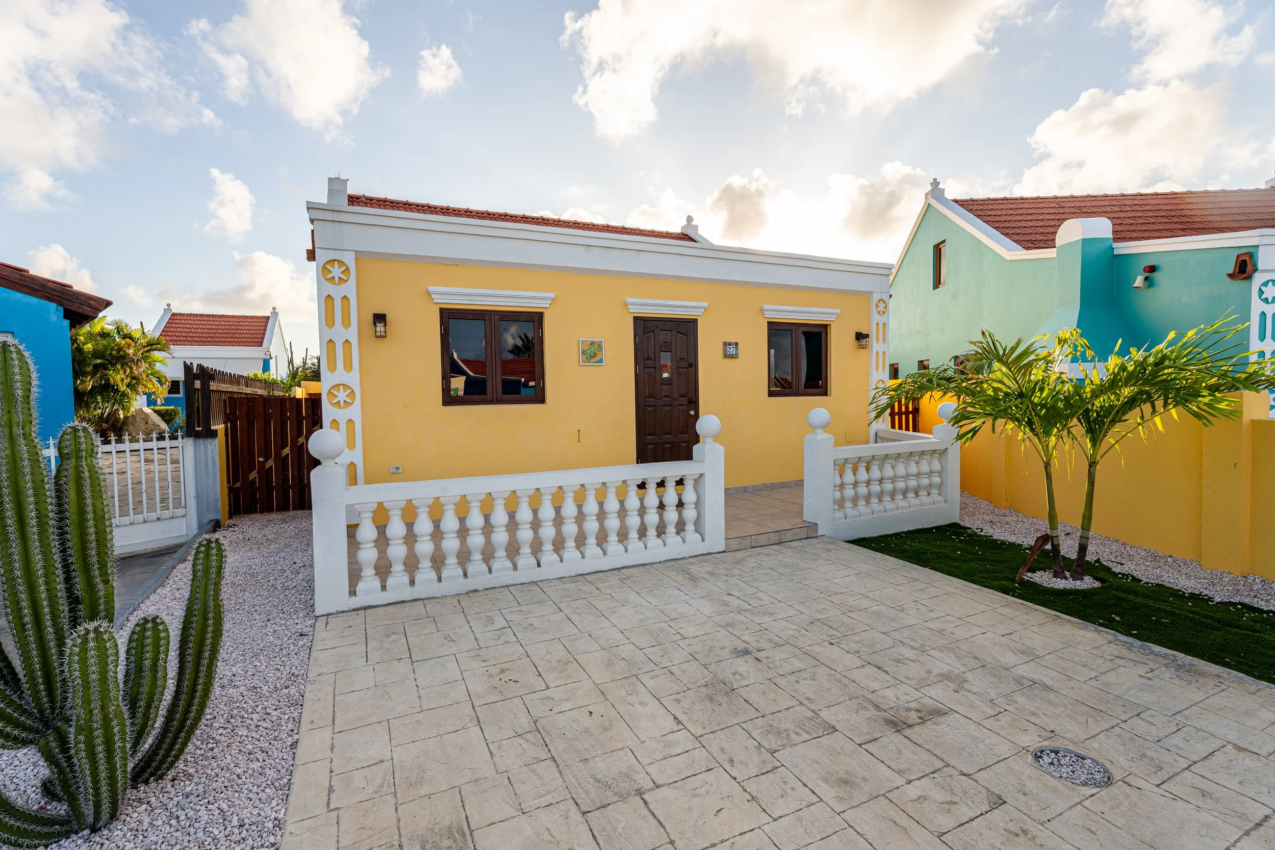Colorful house painted yellow with a white railing, dark brown door, and windows. The front yard has a tiled patio, small palm trees, and cacti, with other vibrant houses visible in the background under a partly cloudy sky.