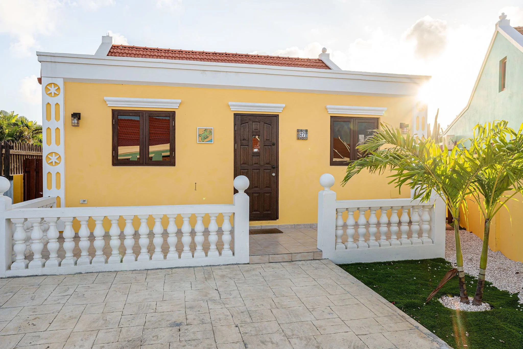 Yellow house with dark brown door and two windows, white decorative fence, small front porch, and a palm tree on the right side, with the sun shining from the right side.