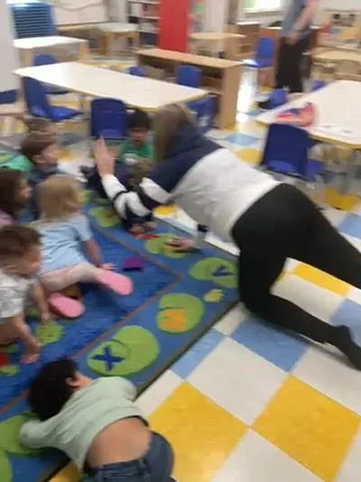A woman is engaging with young children sitting on a colorful rug in a classroom, with some children raising their hands.