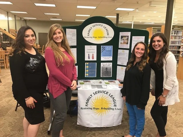 A woman is engaging with young children sitting on a colorful rug in a classroom, with some children raising their hands.