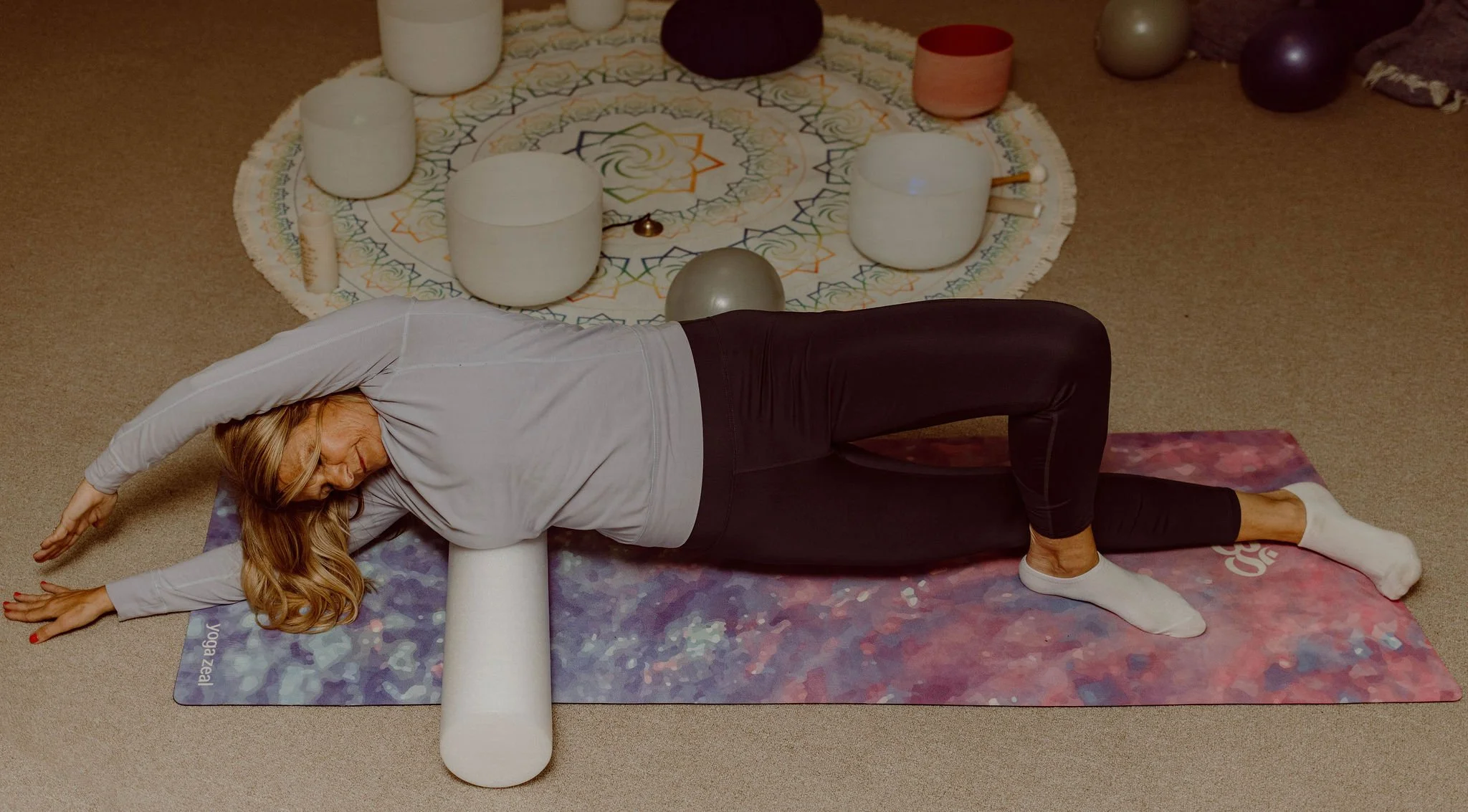 A woman practicing yoga and myofascial release techniques on a yoga mat with a foam roller placed under her neck, in a room that is conductive environment to align the nervous system and support emotional and physucal wellbeing.