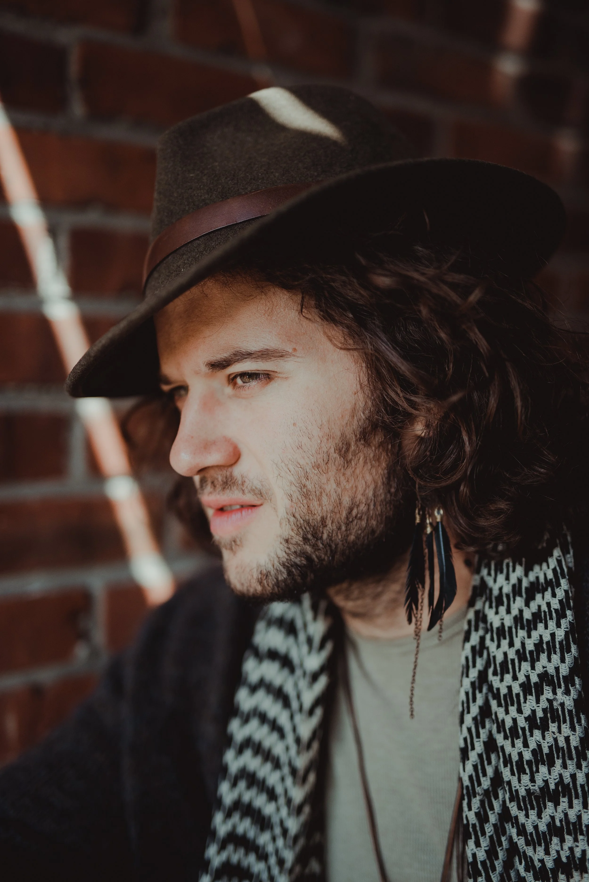A young man with long curly hair wearing a wide-brimmed hat and feather earrings, looking to the side, standing against a brick wall.