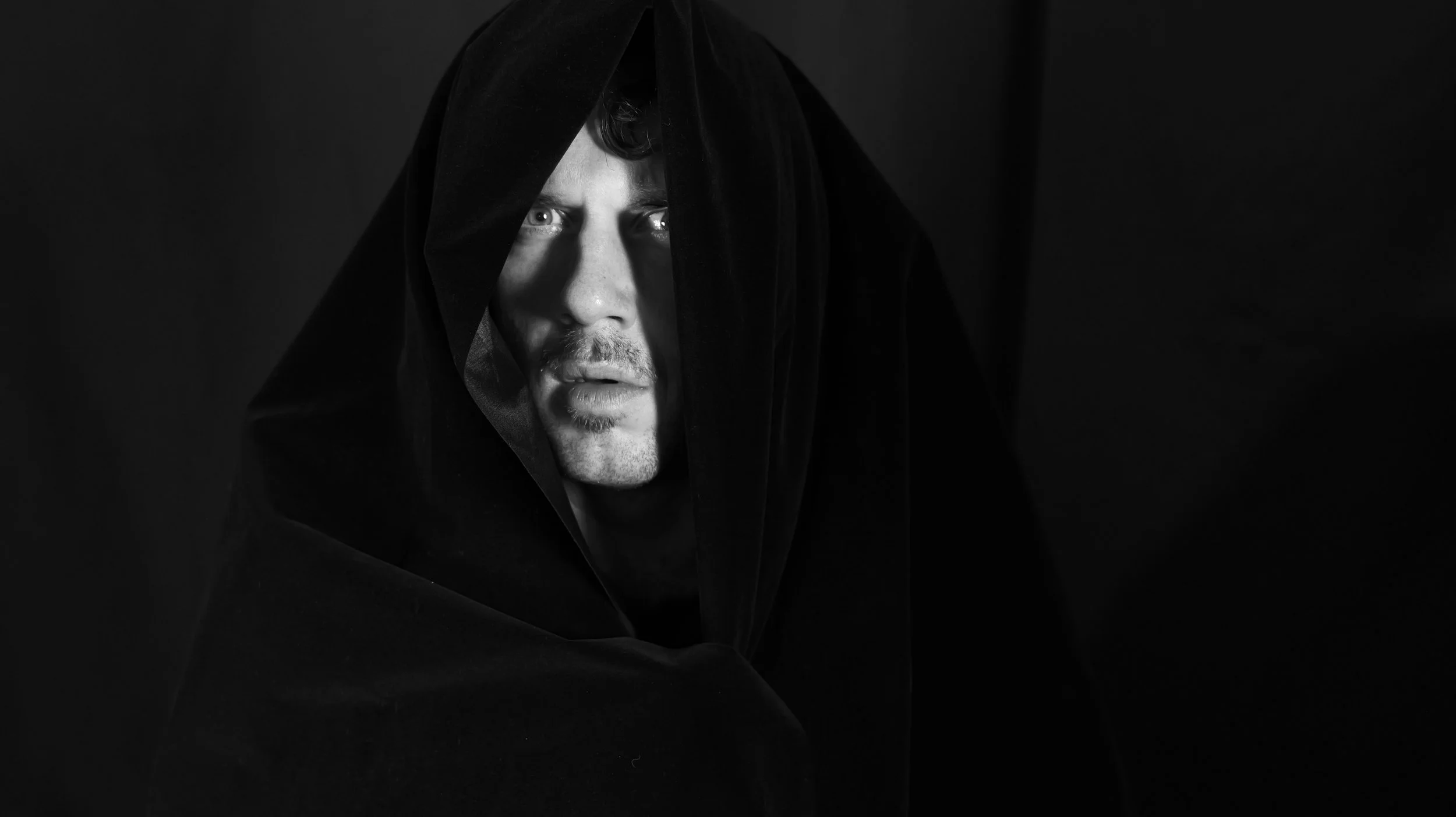 Black and white portrait of a man with a solemn expression, wearing a dark hooded cloak, against a dark background.