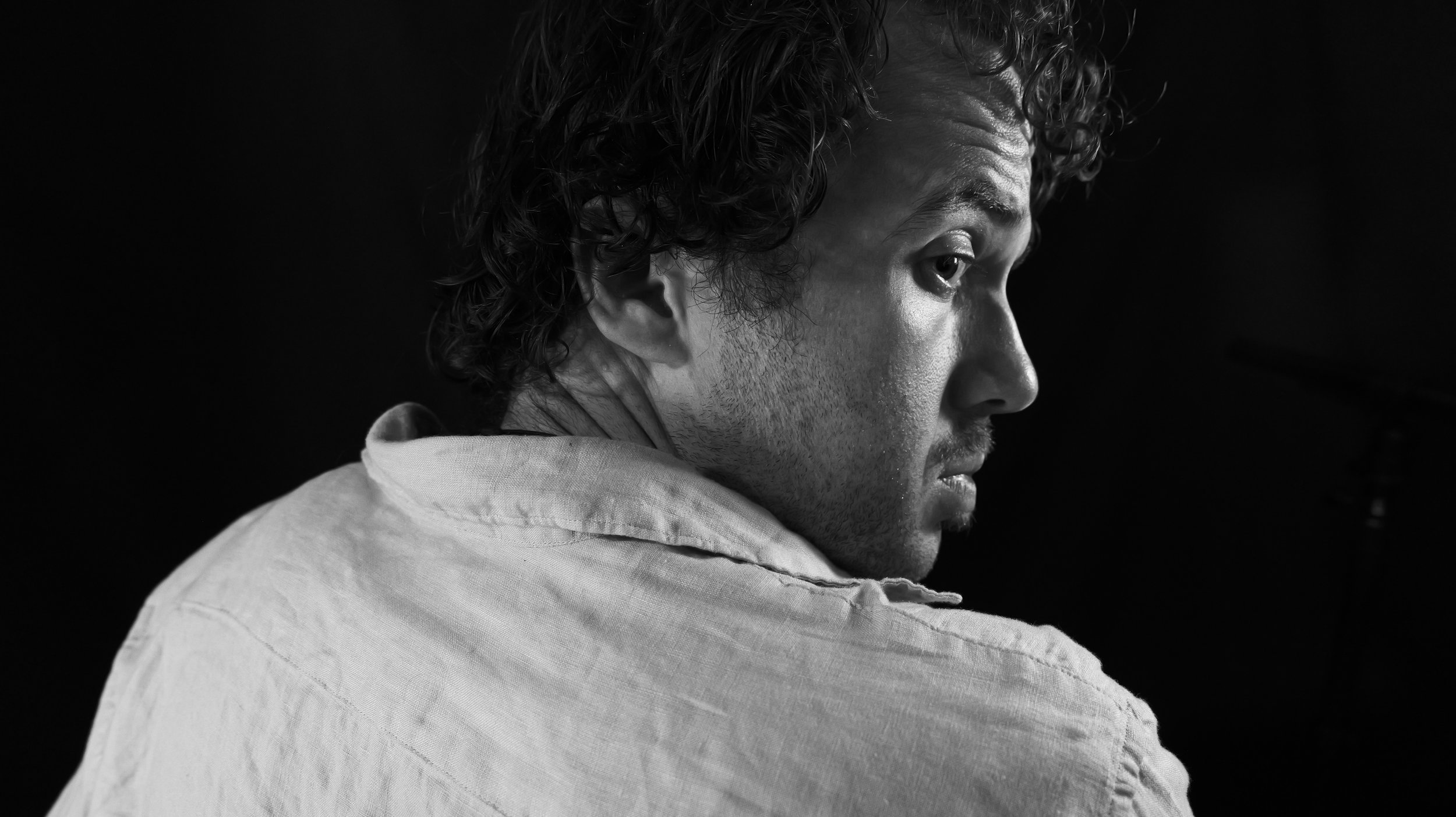 A black and white photo of a man with curly hair, looking over his right shoulder with a contemplative expression, wearing a light-colored shirt with the collar visible.