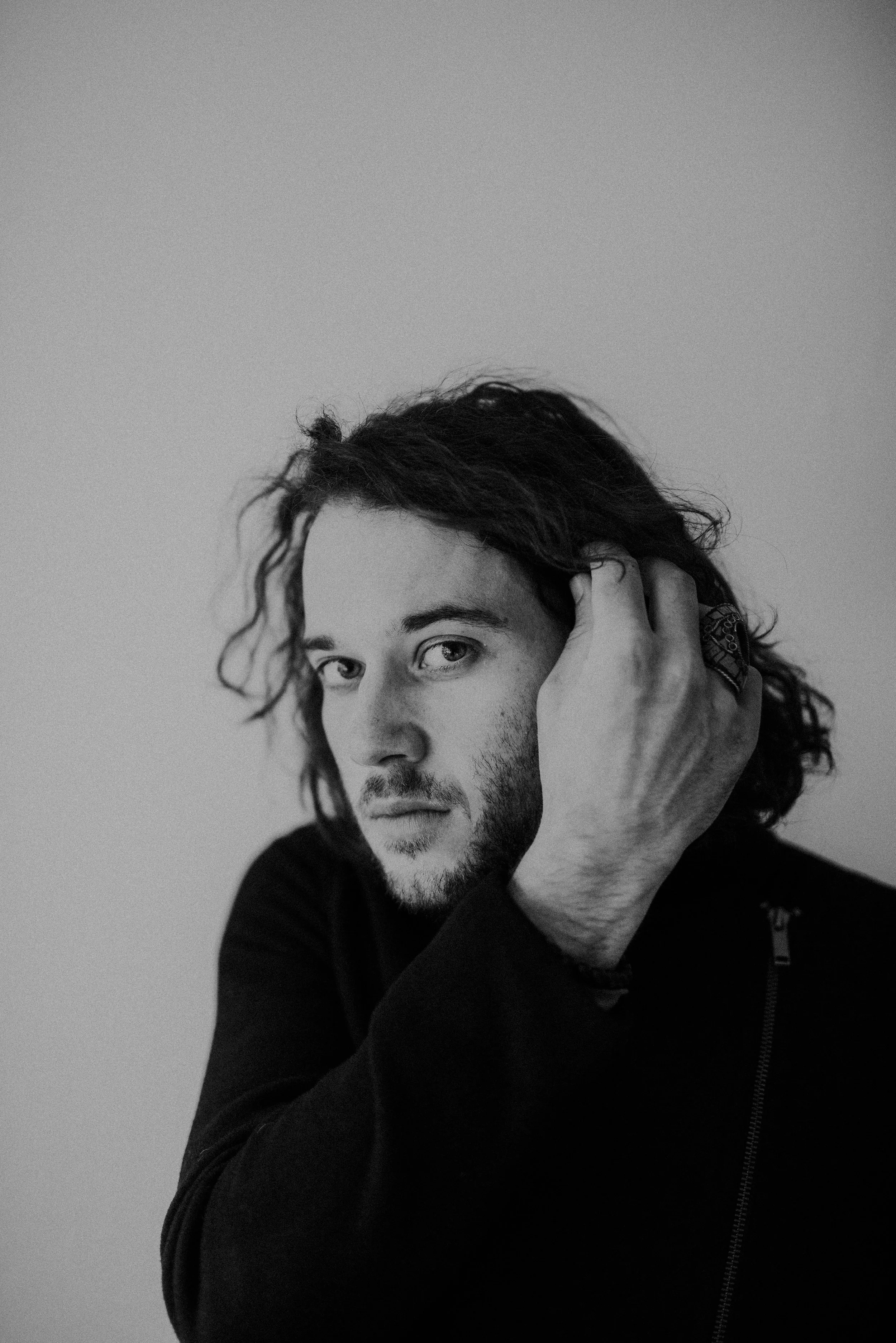 Black and white portrait of a man with long curly hair, a beard, dressed in a dark jacket, looking at the camera with one hand touching his head.