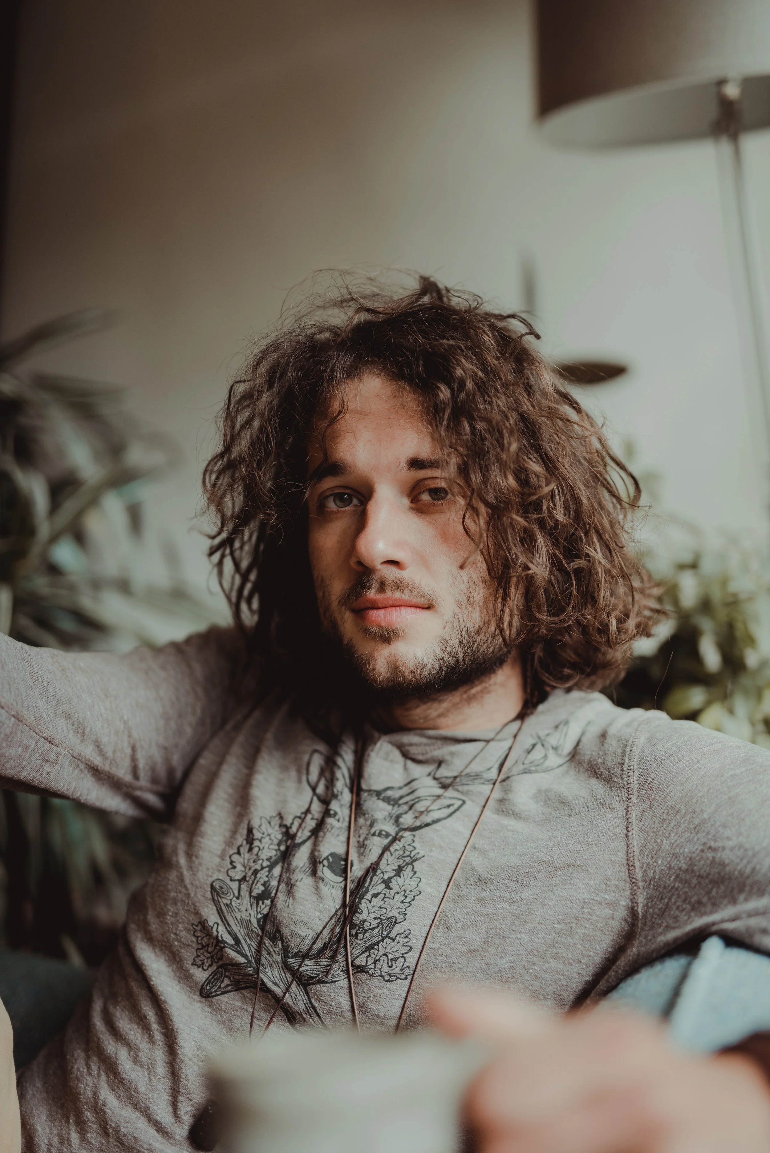 A young man with curly, shoulder-length brown hair and a slight beard, relaxing indoors with one arm behind his head, wearing a light gray sweatshirt with a deer illustration and a necklace, sitting on a sofa with green plants in the background.