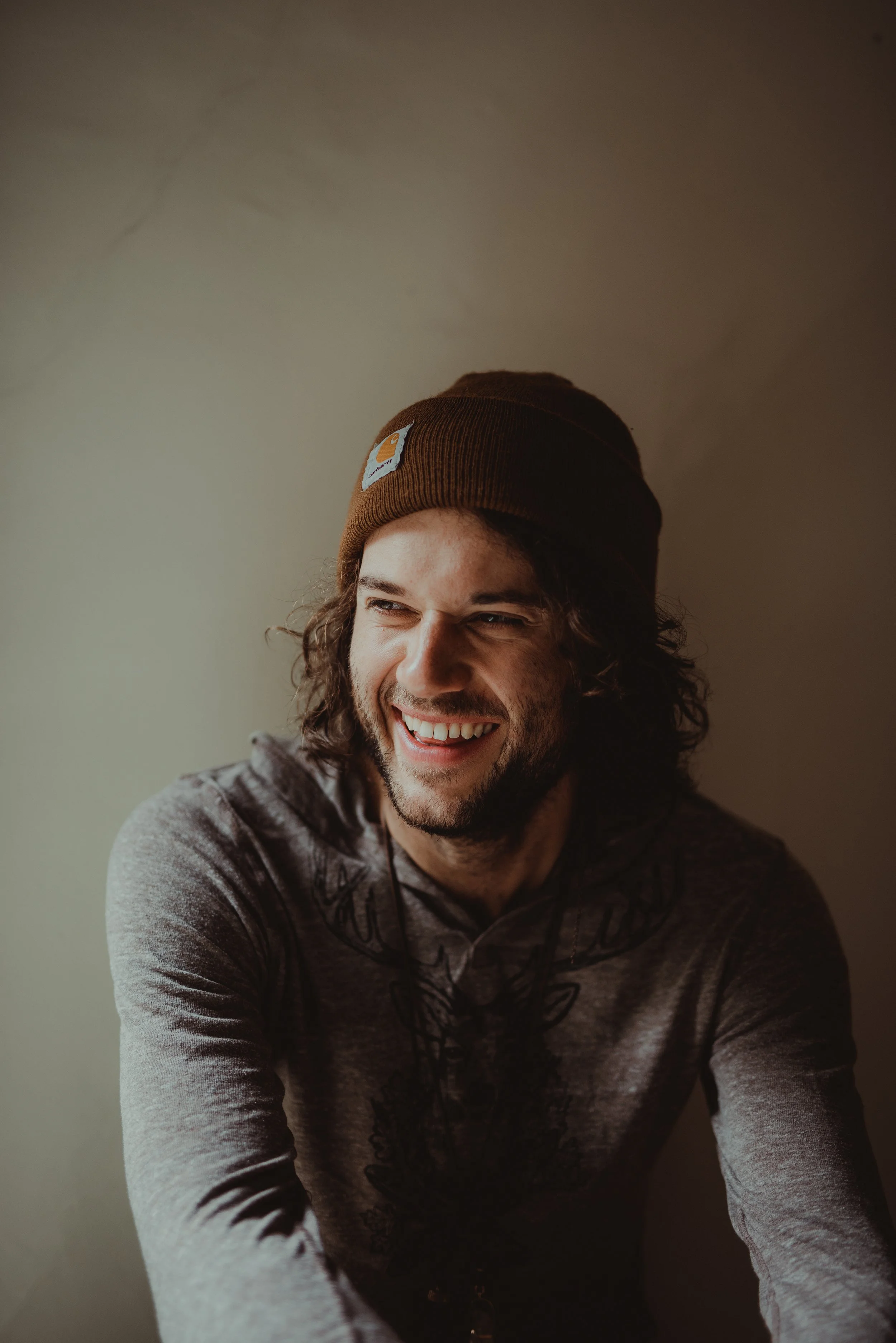 A young man with long dark hair wearing a brown beanie hat and a gray hoodie, smiling and looking off to the side, against a plain background.