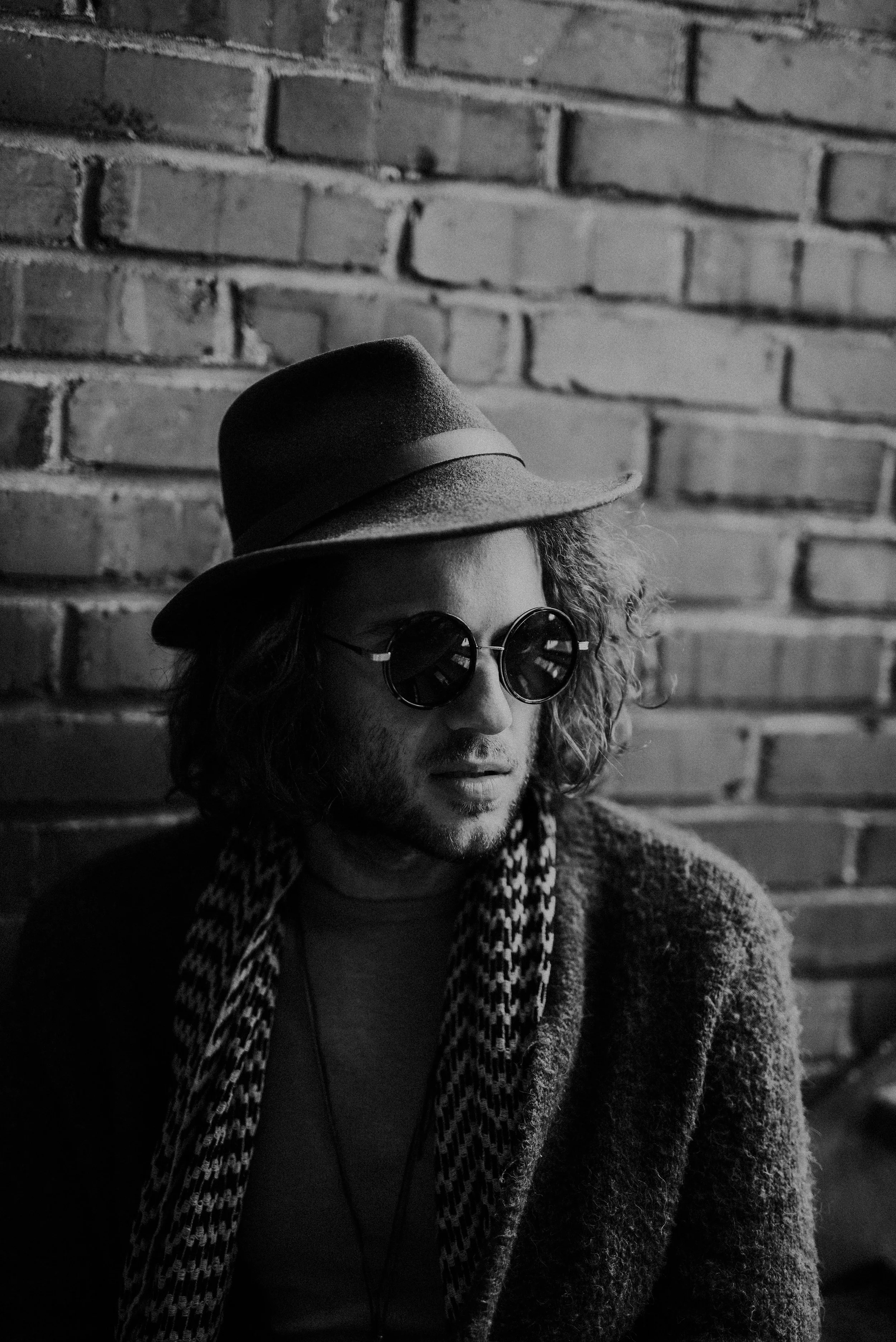A man with curly hair wearing a fedora hat, round sunglasses, and a patterned scarf, sitting against a brick wall in black and white.
