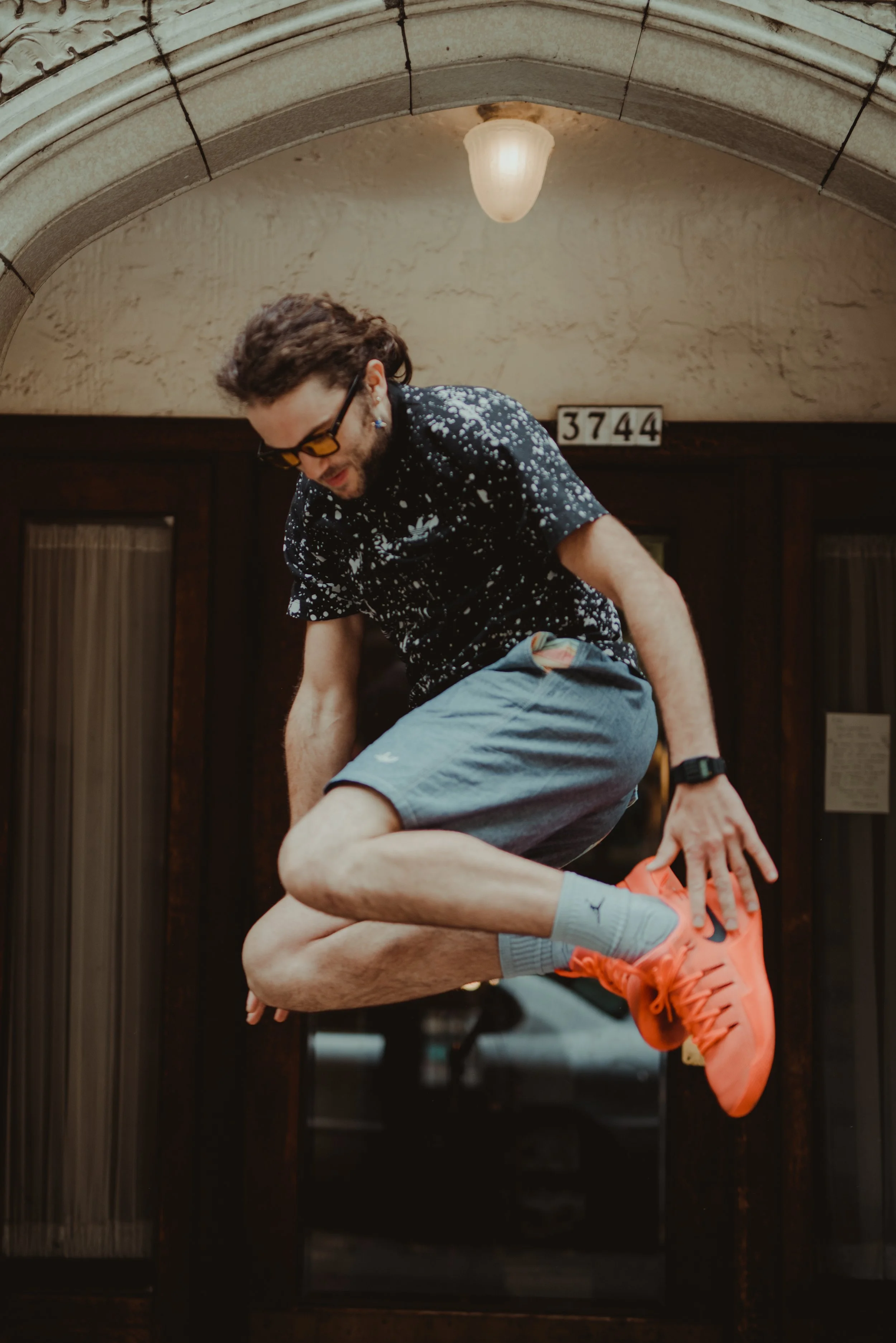 A man jumping in front of a door with the number 3744, wearing a black shirt with white splatters, gray shorts, gray socks with a logo, and bright orange athletic shoes.