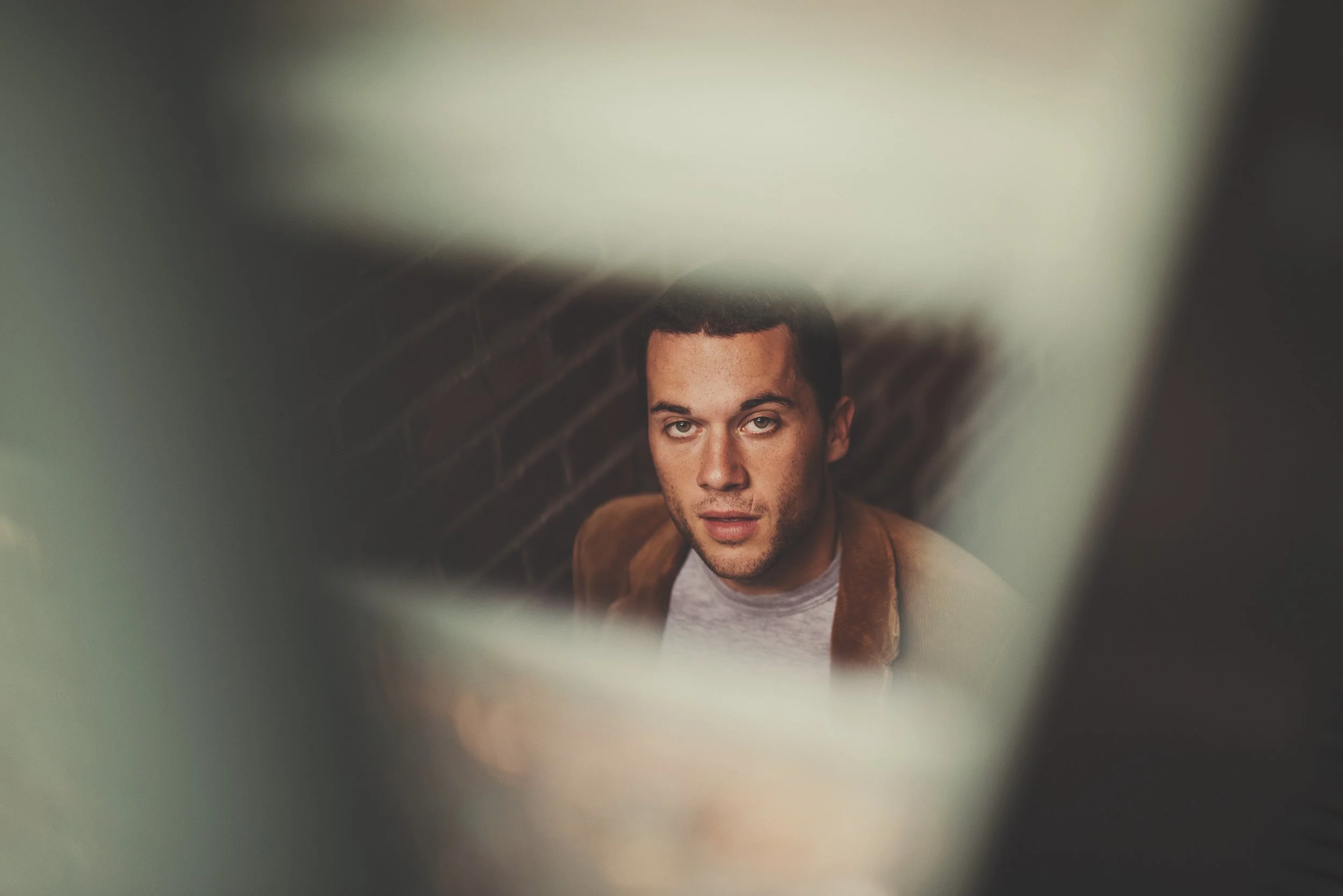 A young man with short dark hair and light skin looks directly up at the camera through an opening, with a brick wall in the background.
