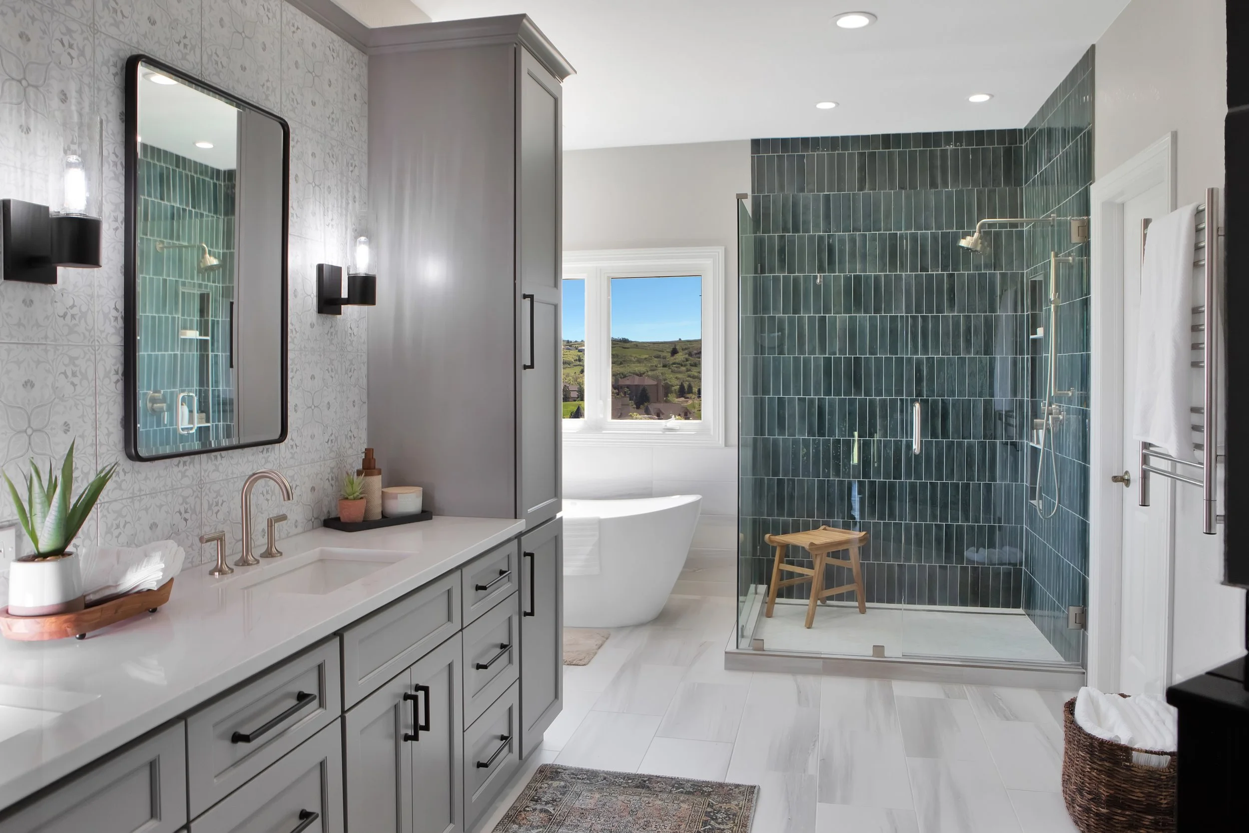 Modern bathroom with gray vanity, white countertop, black hardware, large mirror, black wall sconces, patterned tiles, a freestanding bathtub by window, and a walk-in shower with dark green tiles.
