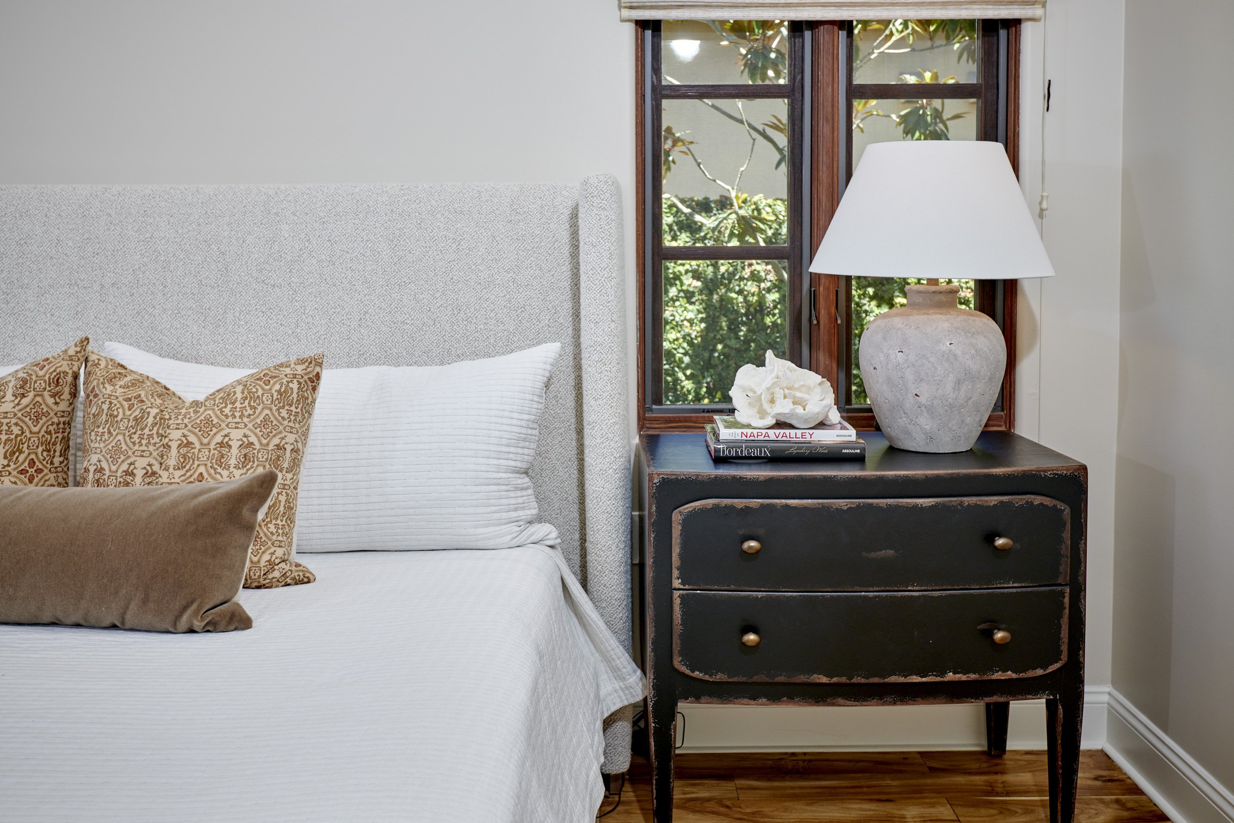 Bedroom with a bed, pillows, and a wooden nightstand with a lamp, books, and decorative items. Window with greenery outside.