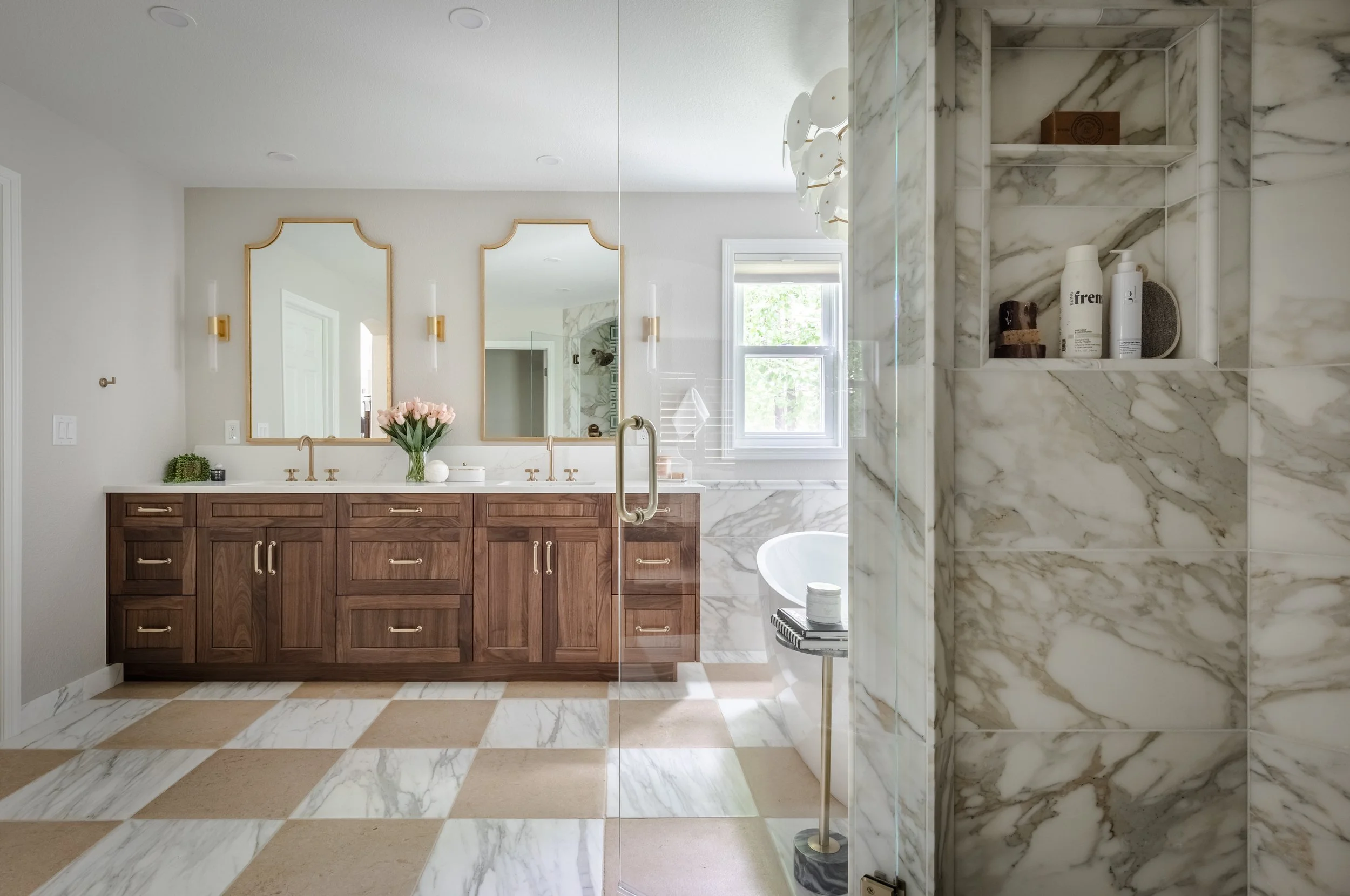 A spacious bathroom with a double vanity featuring a wooden cabinet and two mirrors, a window with natural light, a bathtub in the background, and a glass shower with marble walls in the foreground.