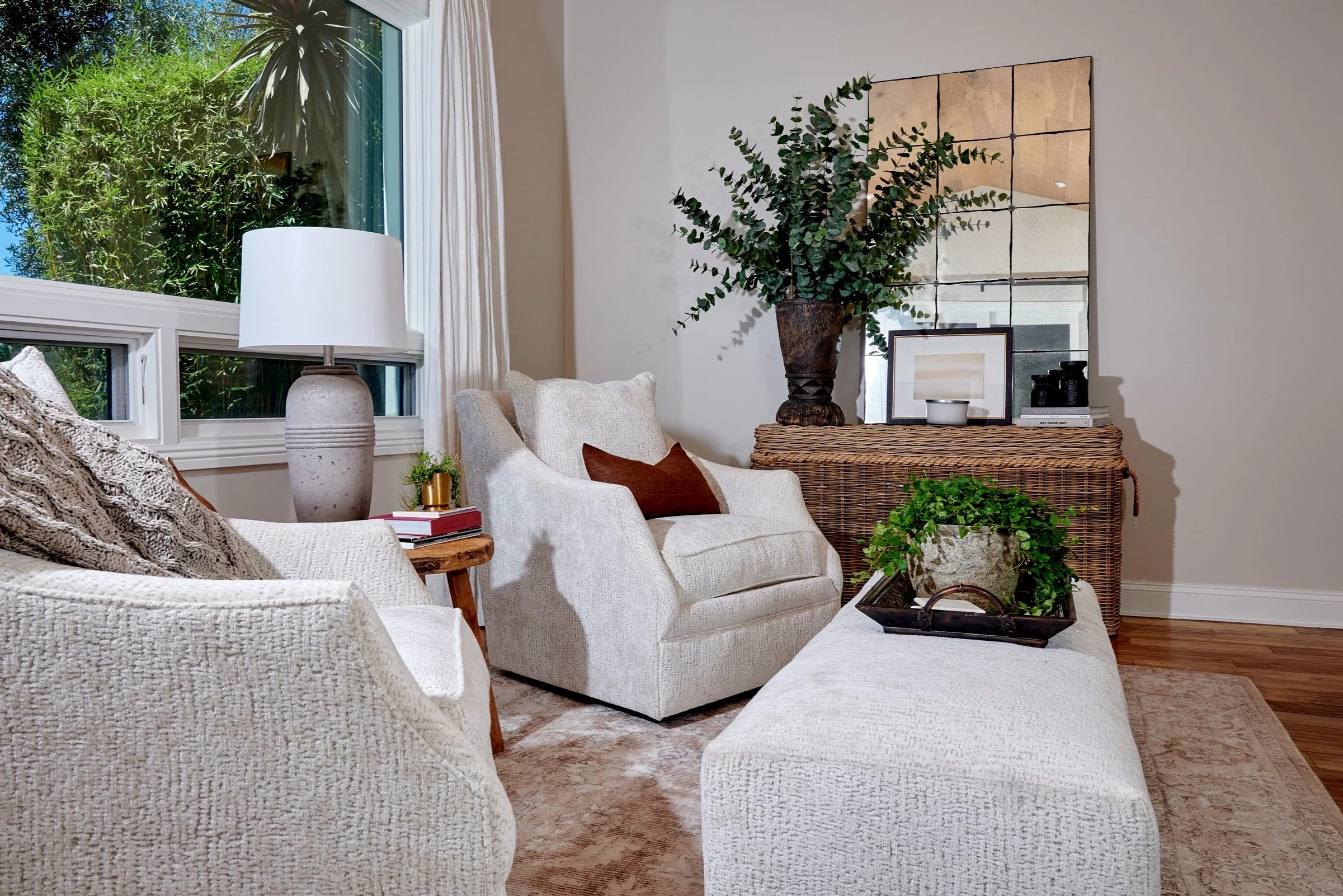 Living room with beige upholstered chairs, a wooden side table with books and a small plant, large window with white curtains, a wicker console table with a large plant, a framed picture, and decorative items, a mirror on the wall, and a beige area rug.
