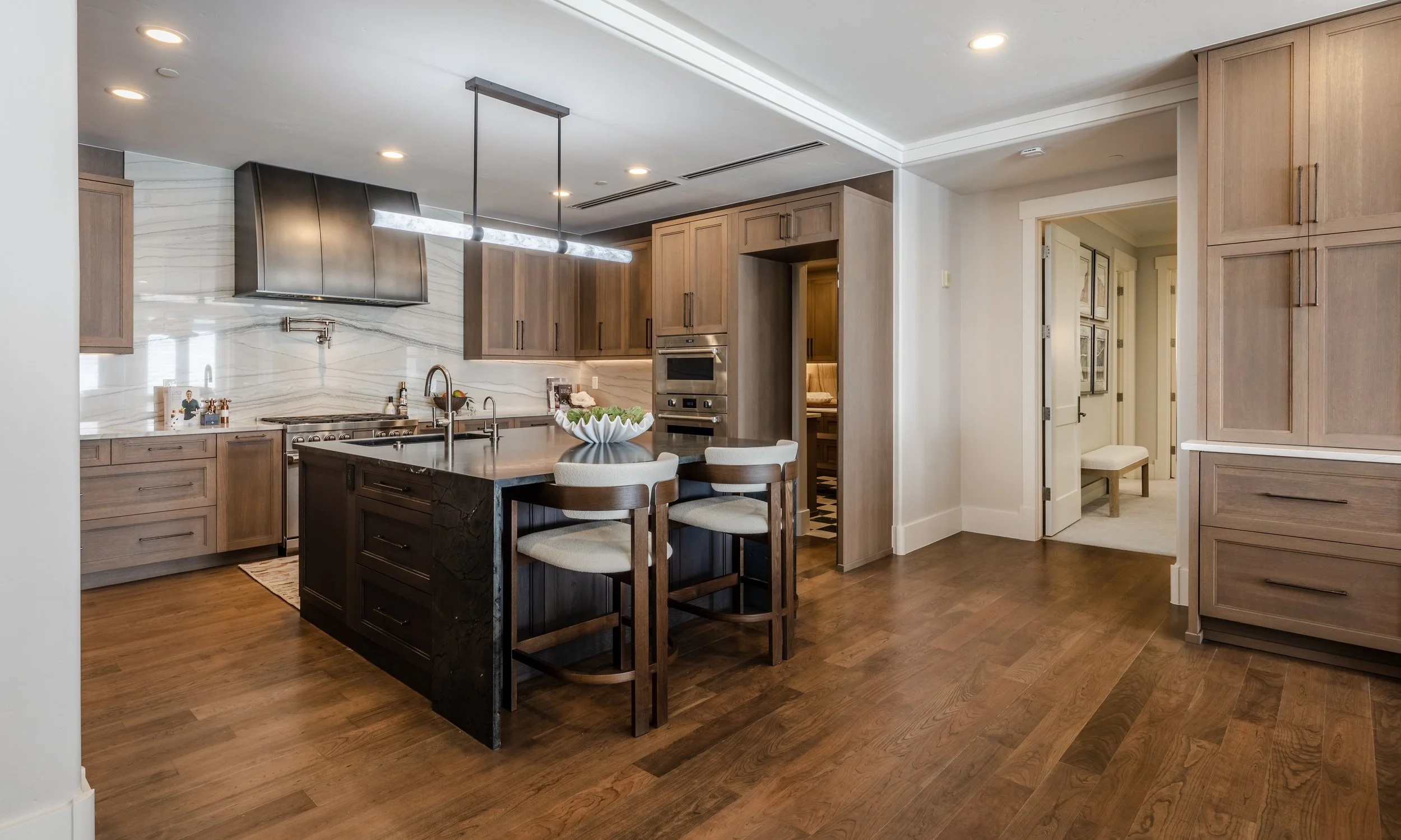 Modern kitchen with wooden cabinets, a black kitchen island, stainless steel appliances, a marble backsplash, and a dining area with four chairs. Hardwood floors and recessed ceiling lights.