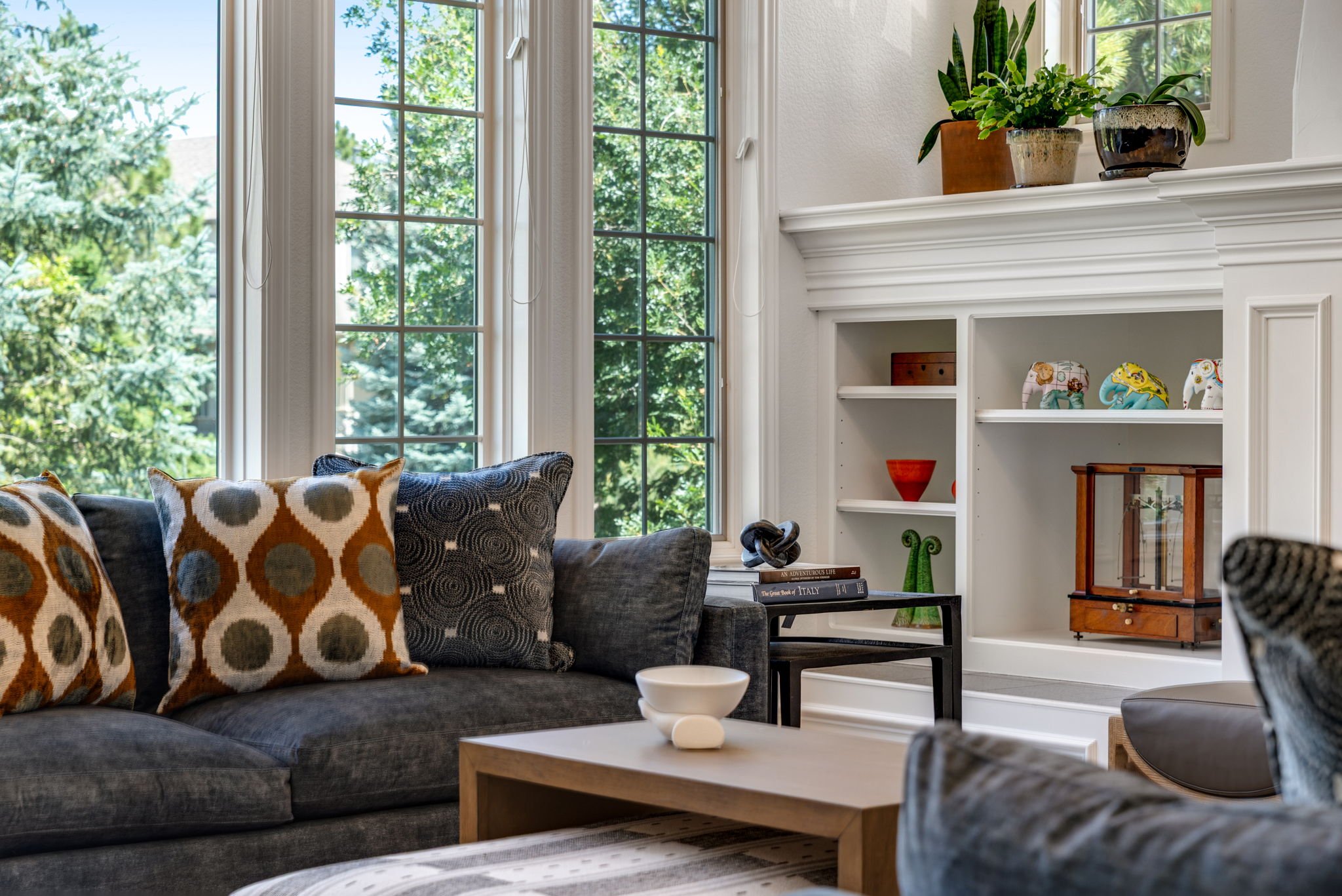 Living room with large windows, dark gray sofa with patterned pillows, white built-in bookcase with decorative items, and potted plants on top of the bookcase.