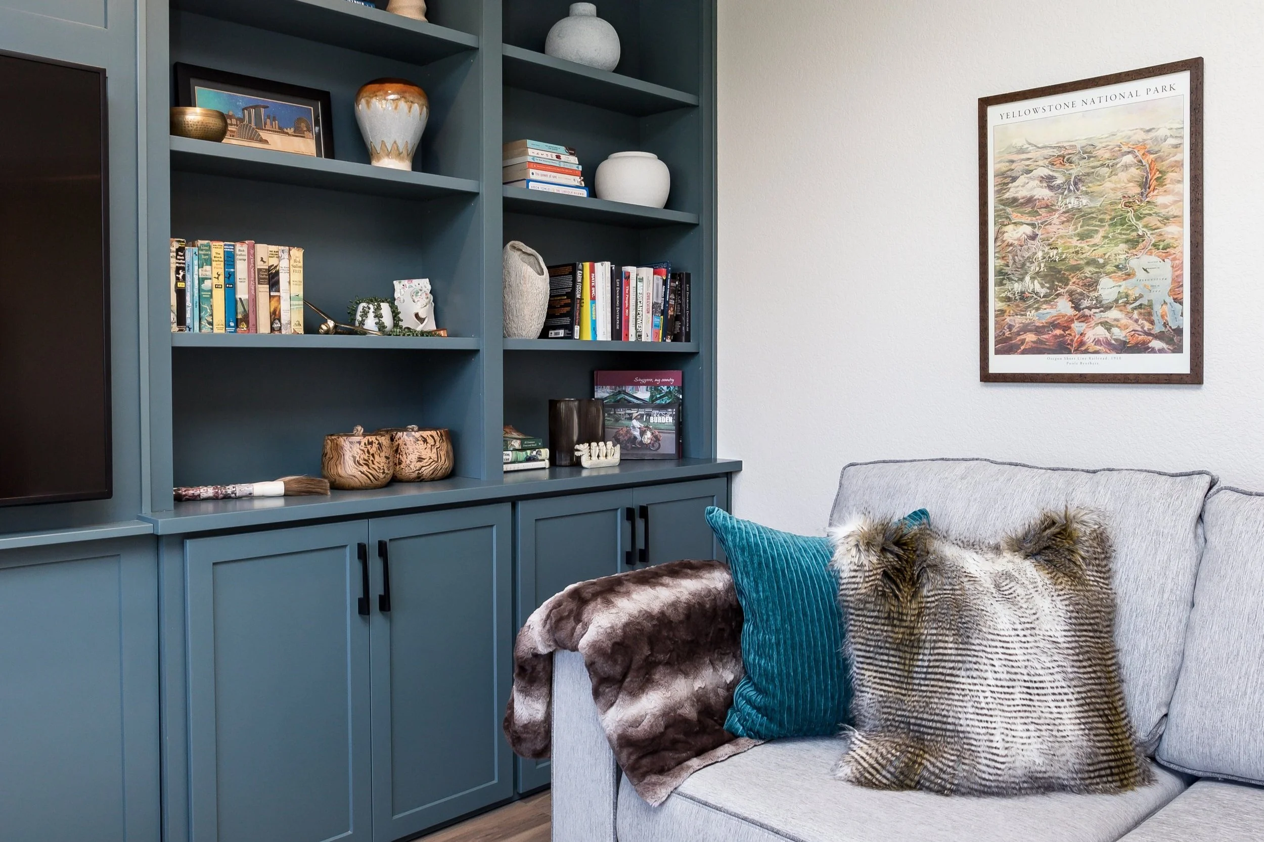 Living room with a teal bookshelf filled with books and decorative items, a framed Yellowstone National Park map on the wall, and a grey sofa with colorful pillows and a faux fur throw.