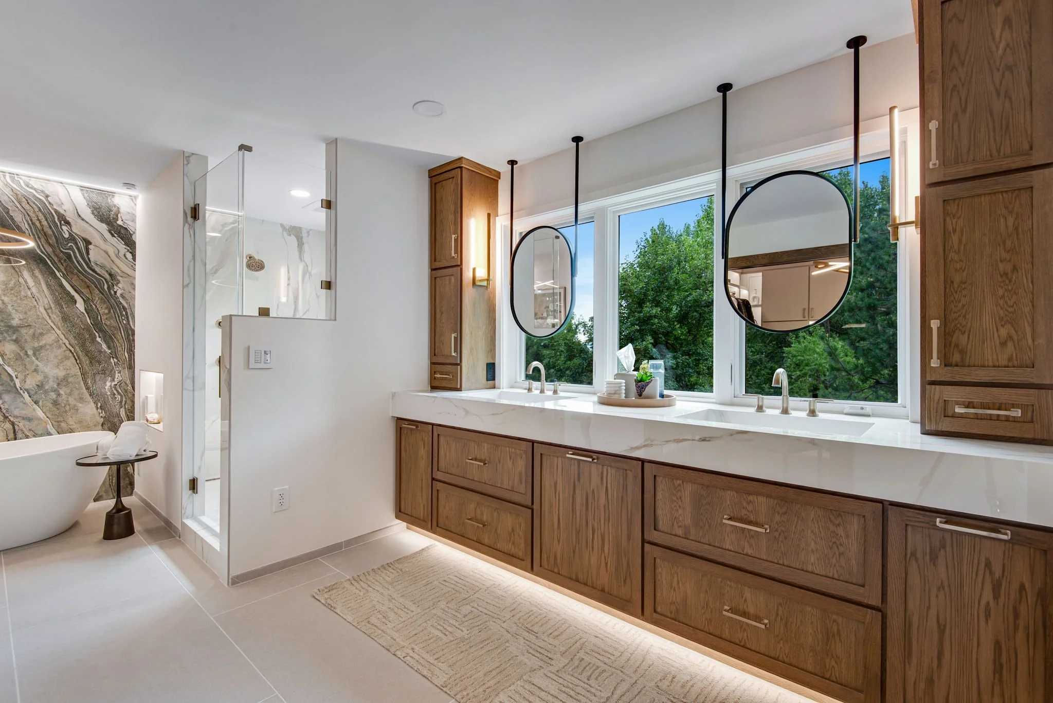 Modern bathroom with large window, wooden cabinetry, oval mirrors, and a marble countertop. A bathtub with a floor-mounted faucet and a towel rack are visible in the corner.