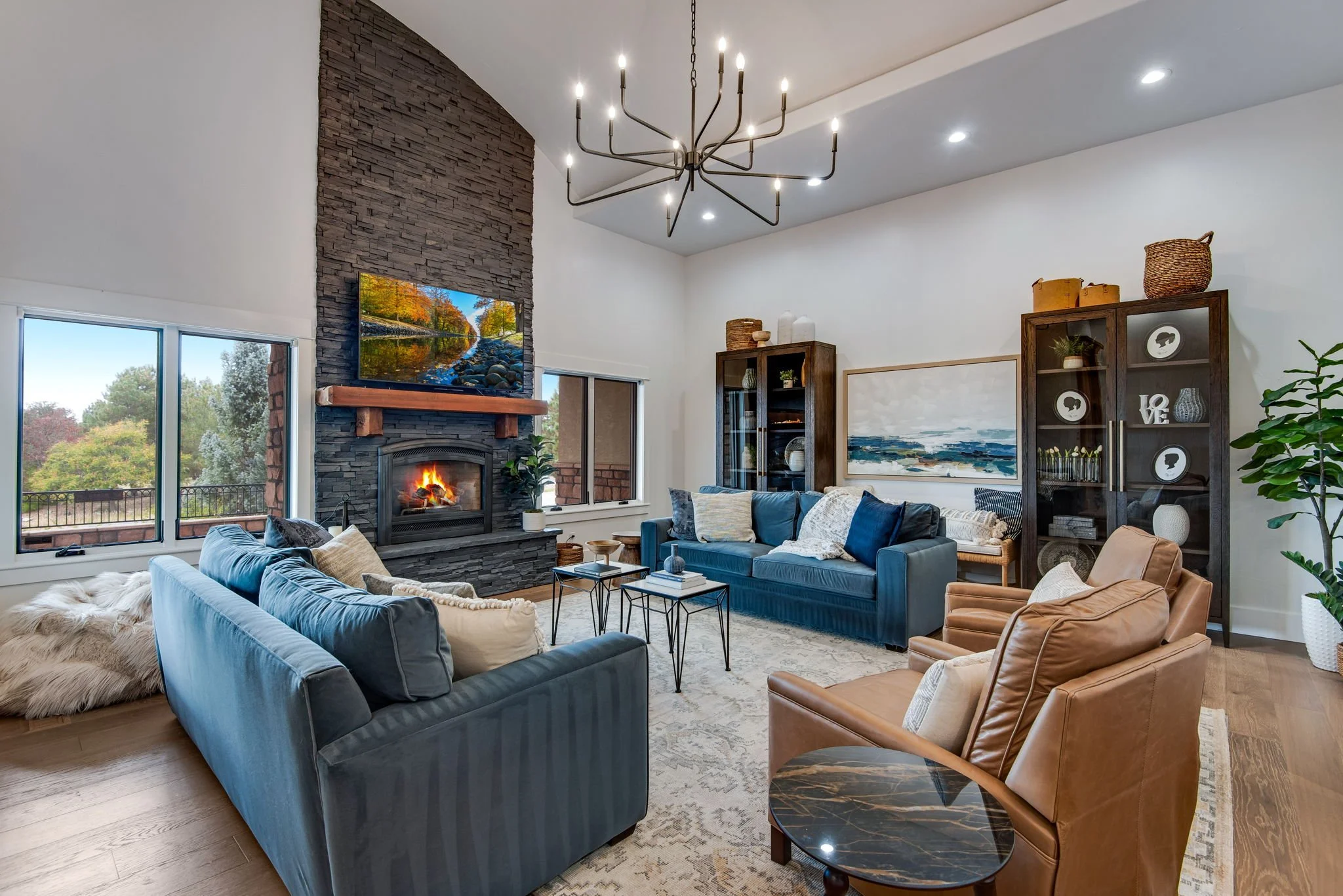 Living room with blue and tan sofas, a black stone fireplace with a fire, wall-mounted TV, large windows, wooden shelves, a modern chandelier, and decorative items.