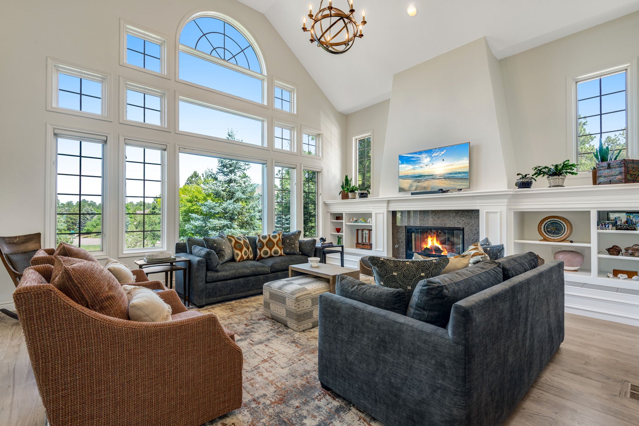 Living room with large windows, a fireplace with a fire, a TV above the fireplace, and various sofas and chairs around a coffee table, with decorative items and plants on the shelves and windowsills.